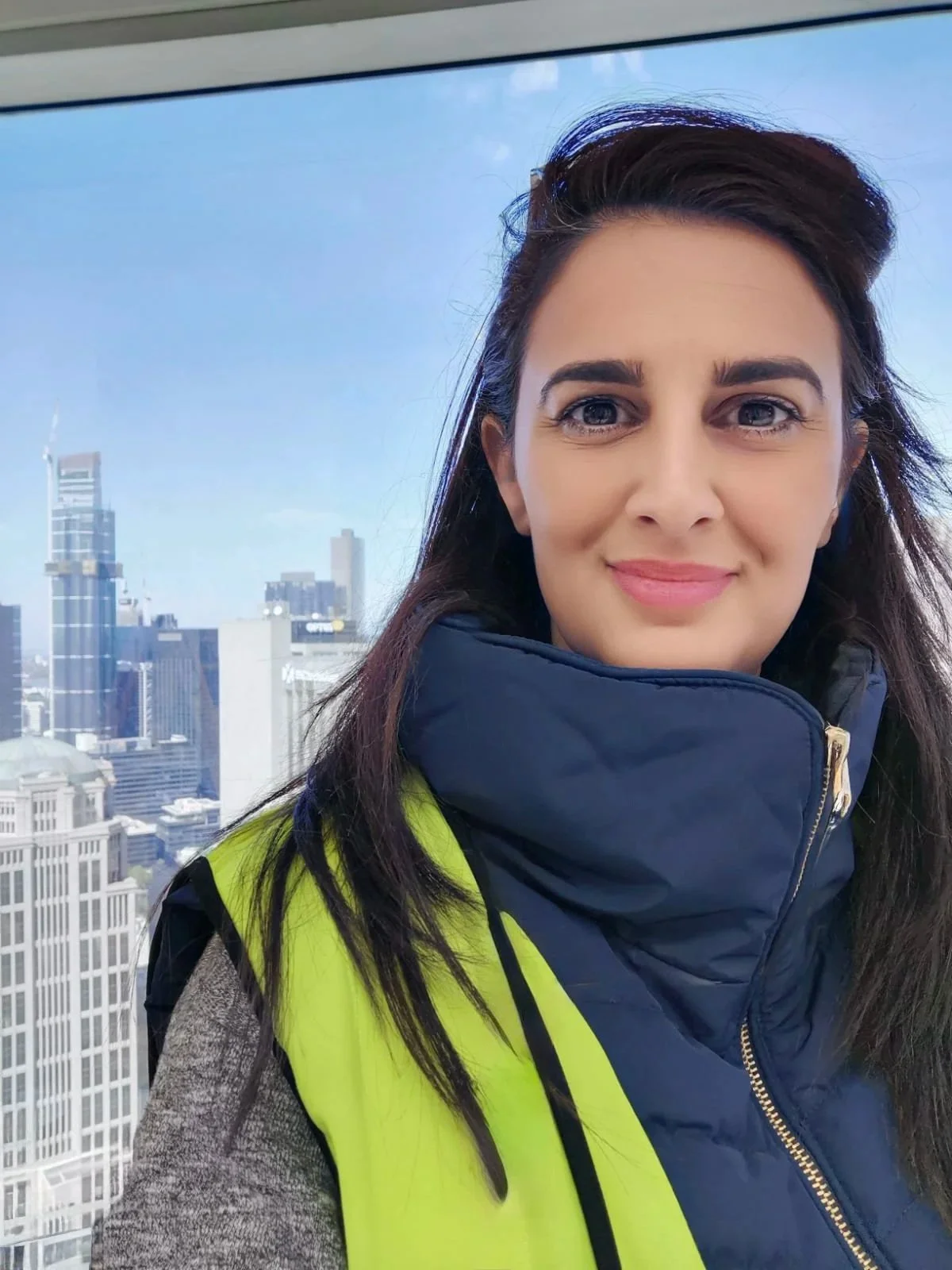 A woman taking a selfie with a city skyline behind her, wearing a high-visibility safety vest and a navy jacket.