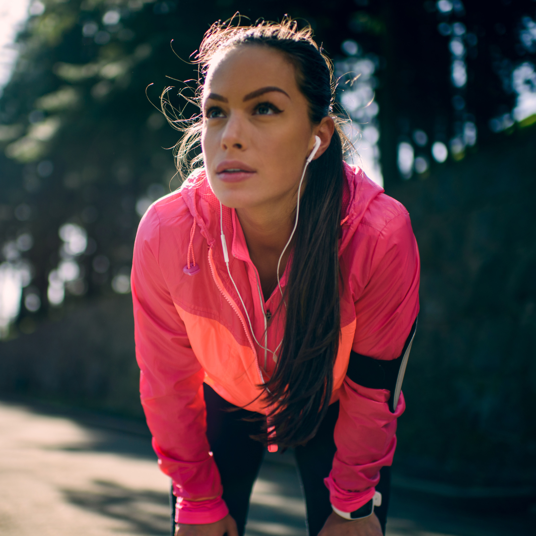 A woman in a pink athletic jacket and black leggings is outdoors, wearing earbuds, bending slightly forward, likely after exercise.
