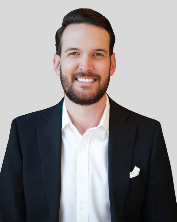 Professional portrait of a man with dark hair and a beard, wearing a black blazer and white shirt, smiling at the camera.