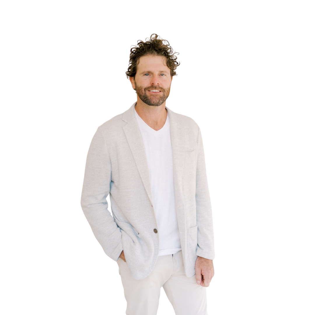 A man with curly brown hair and a beard standing against a white background, wearing a light-colored blazer, white t-shirt, and light-colored pants, smiling and looking at the camera.