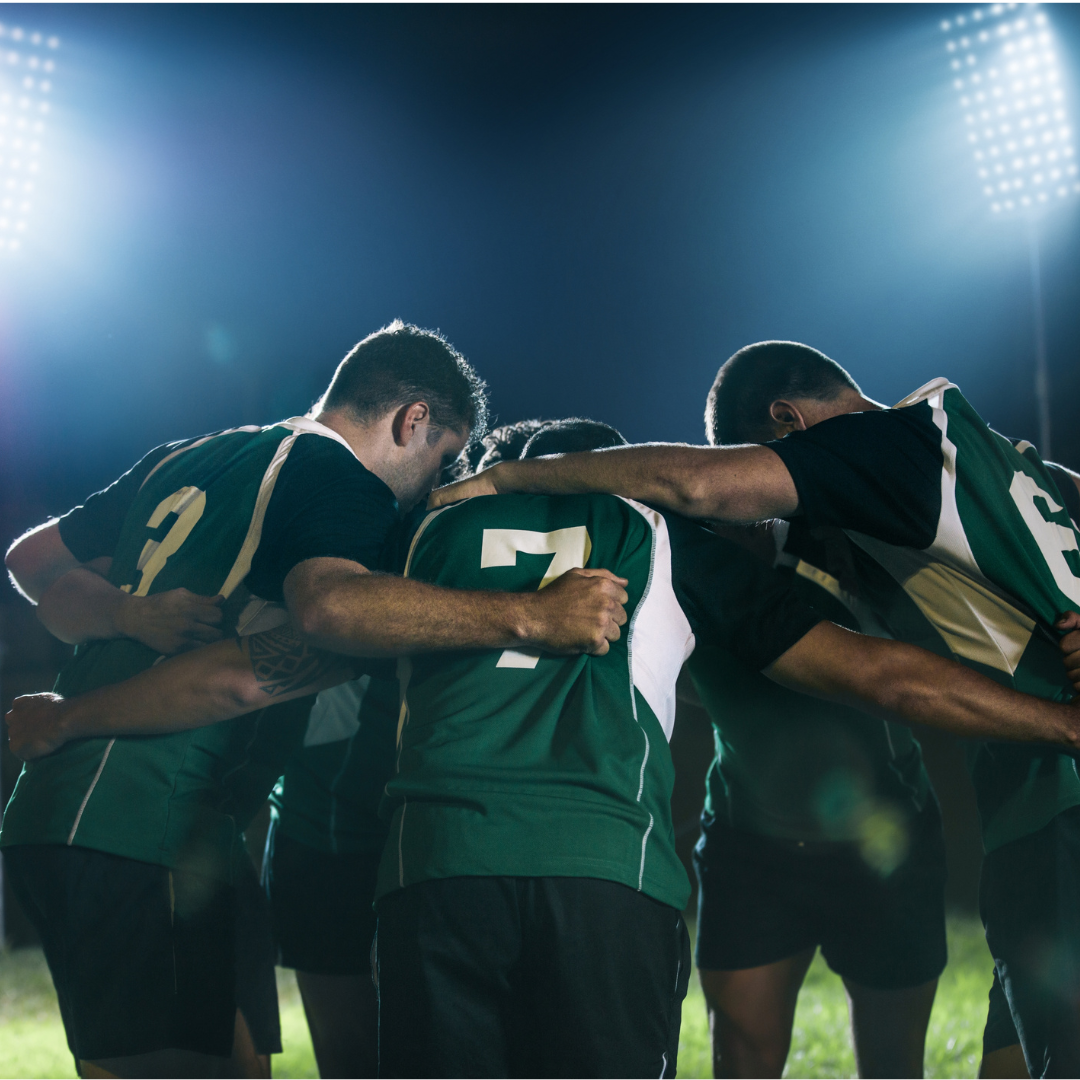 A rugby team huddles together on the field during a game at night, illuminated by bright stadium lights.