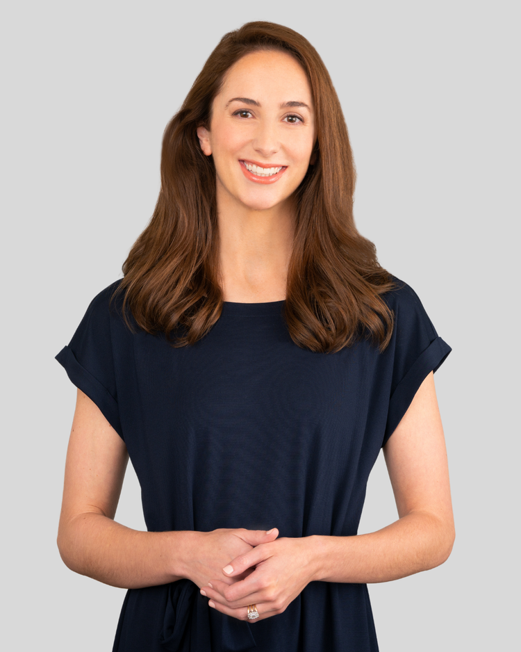 A woman with long, wavy brown hair smiling, wearing a navy blue dress, standing against a light gray background.