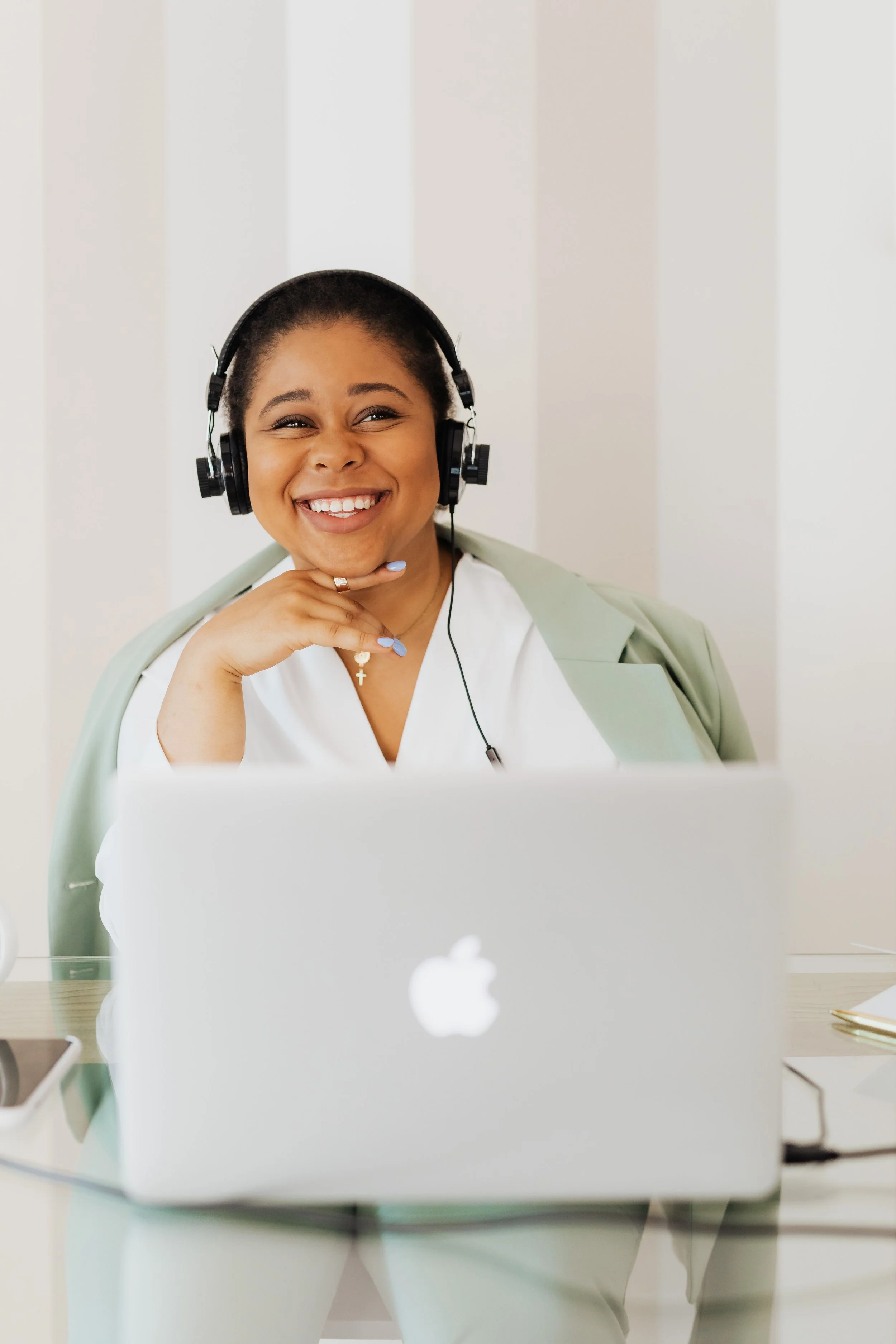 A woman with a big smile, wearing headphones, sitting in front of a laptop, with a smartphone on the table, in a bright room with a striped wall.