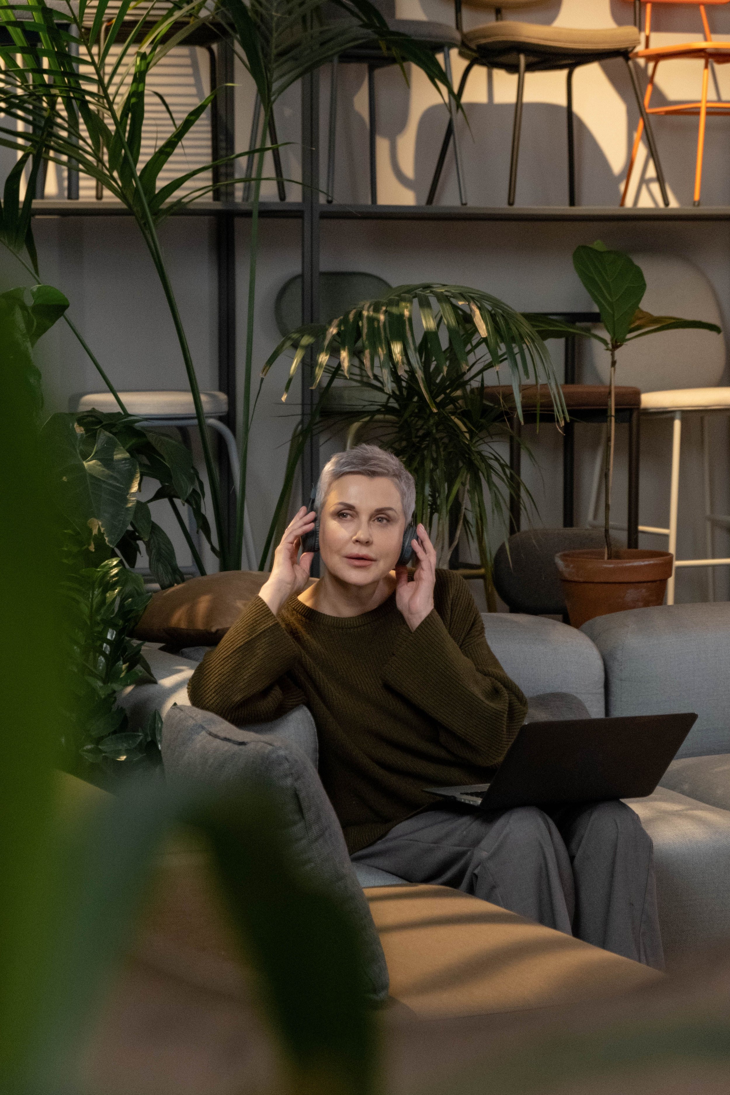A woman with short gray hair wearing a brown sweater sitting on a sofa with a laptop on her lap, surrounded by green potted plants and chairs on a shelf behind her.