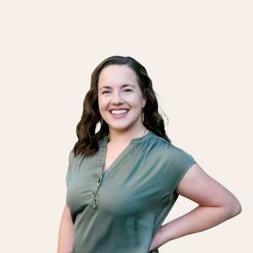 A woman with shoulder-length wavy brown hair, smiling, wearing a teal short-sleeve top, standing against a plain light background.