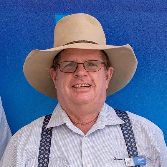A smiling man wearing glasses, a wide-brimmed tan hat, a light-colored striped shirt, and suspenders, standing in front of a bright blue wall.
