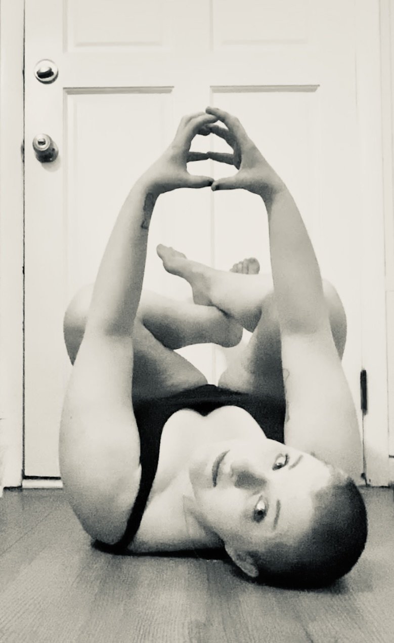 woods is on a wood floor with a white paneled door behind them. they are laying on their back with their knees to their chest, their feet crossed, their arms up in front of them with their hands closed together to make a circle. their head is twisted