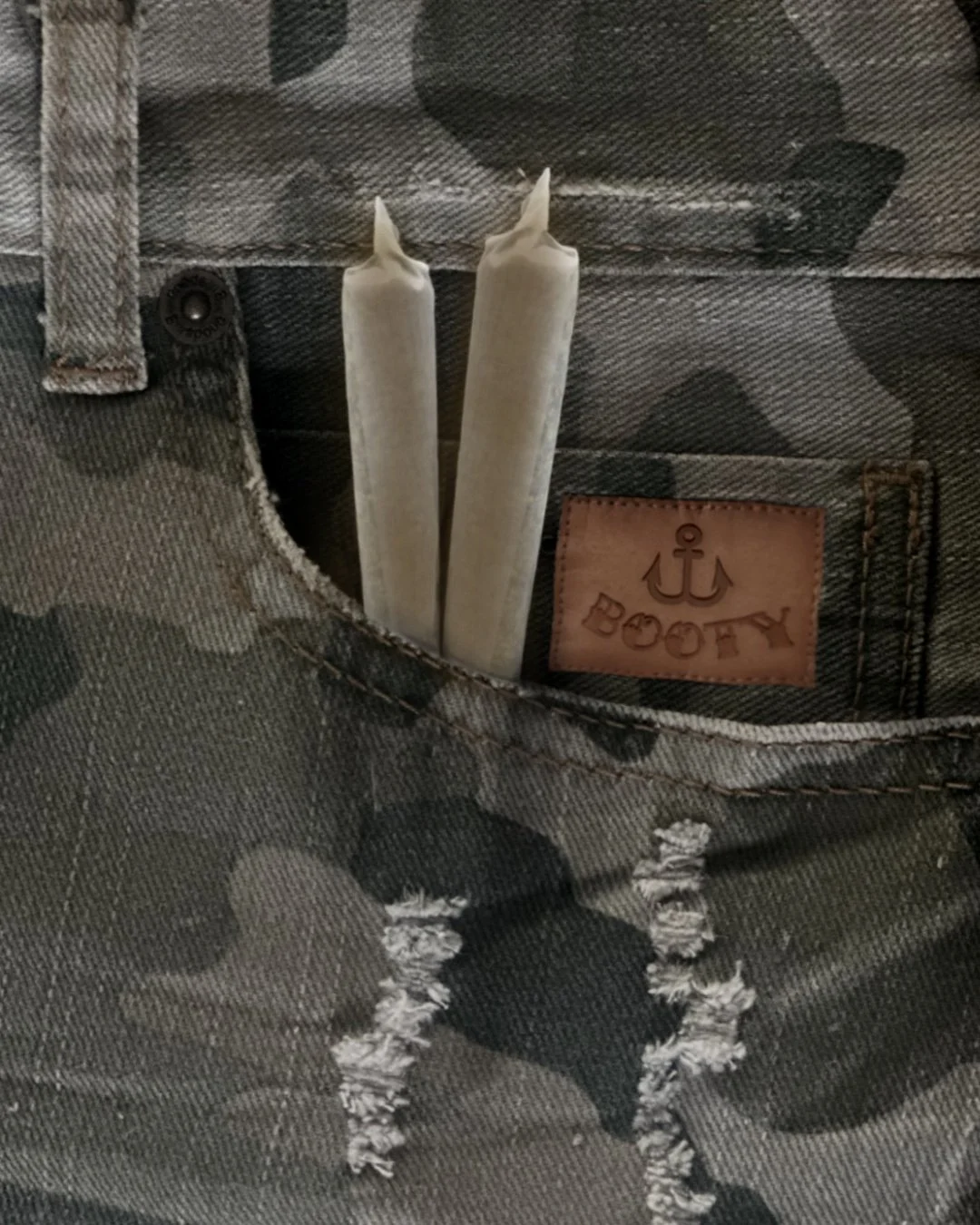 Camouflage patterned pants with a leather patch that says 'BOOY' and an anchor symbol, with two white candles in the front pocket.