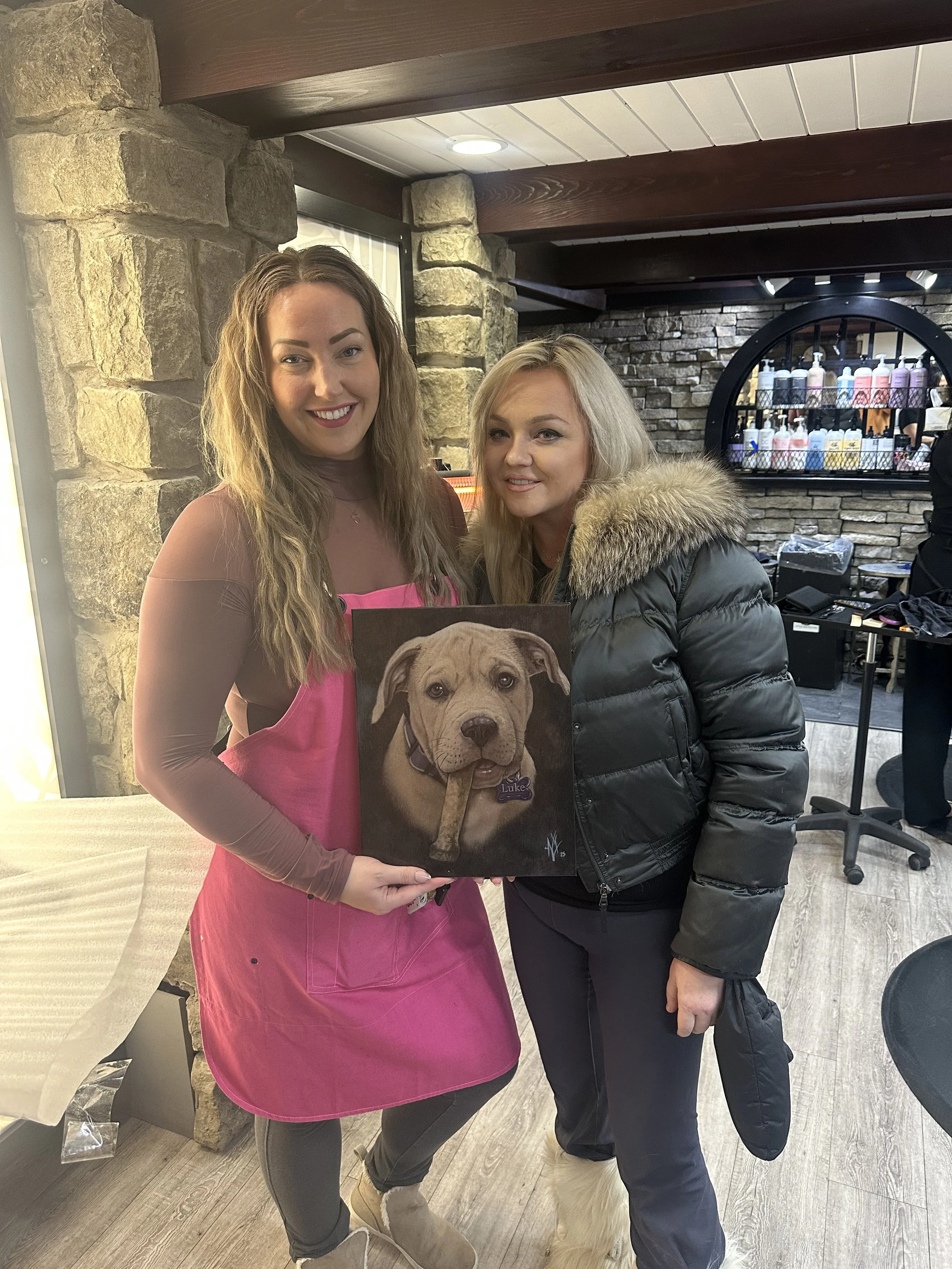 Two women standing together, one wearing a pink apron and holding a painting of a dog with a bone in its mouth, inside a room with stone walls and a bar in the background.