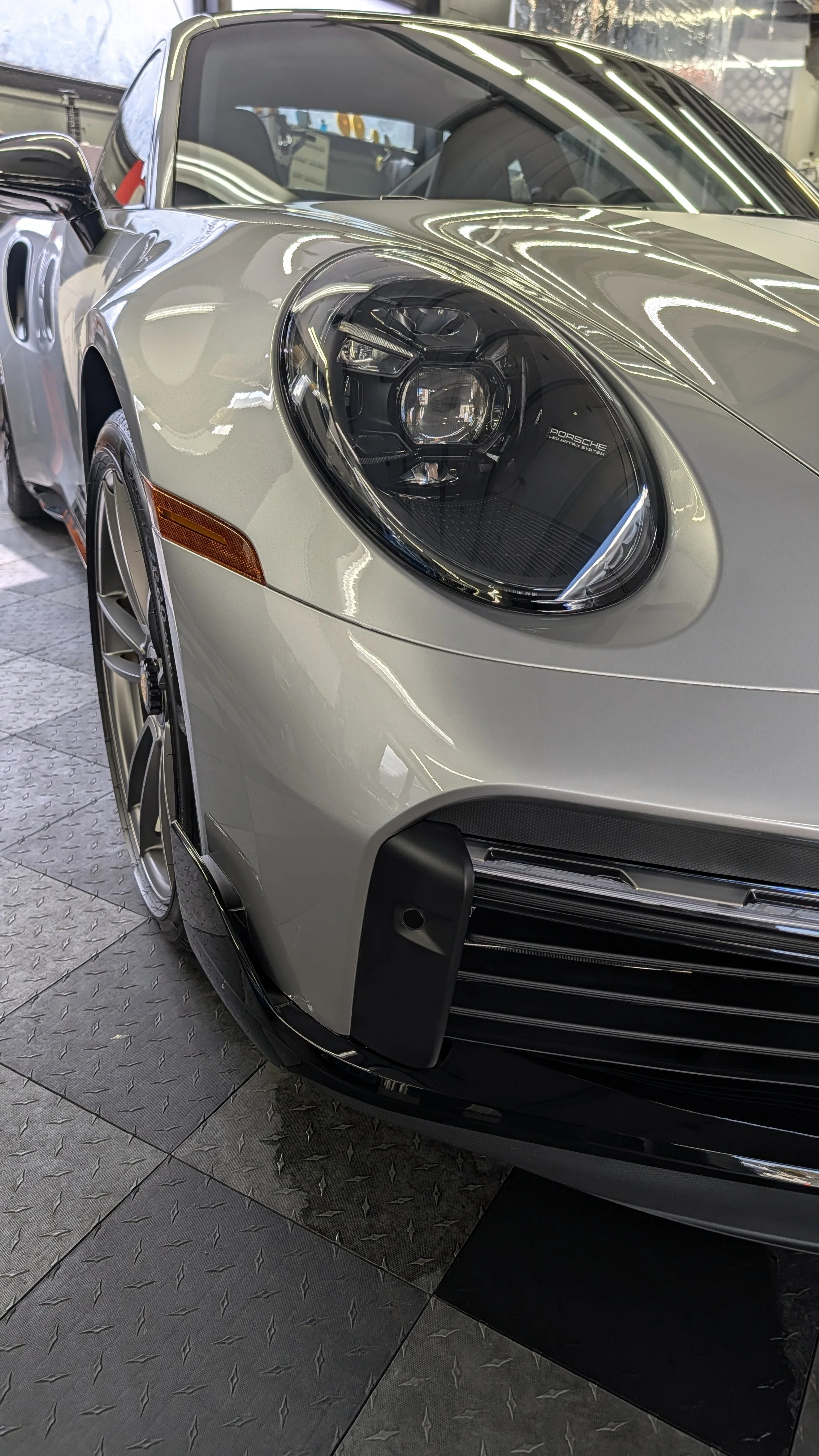 Close-up view of the front end of a silver Porsche car inside a showroom, highlighting the headlight, front bumper, and part of the wheel.