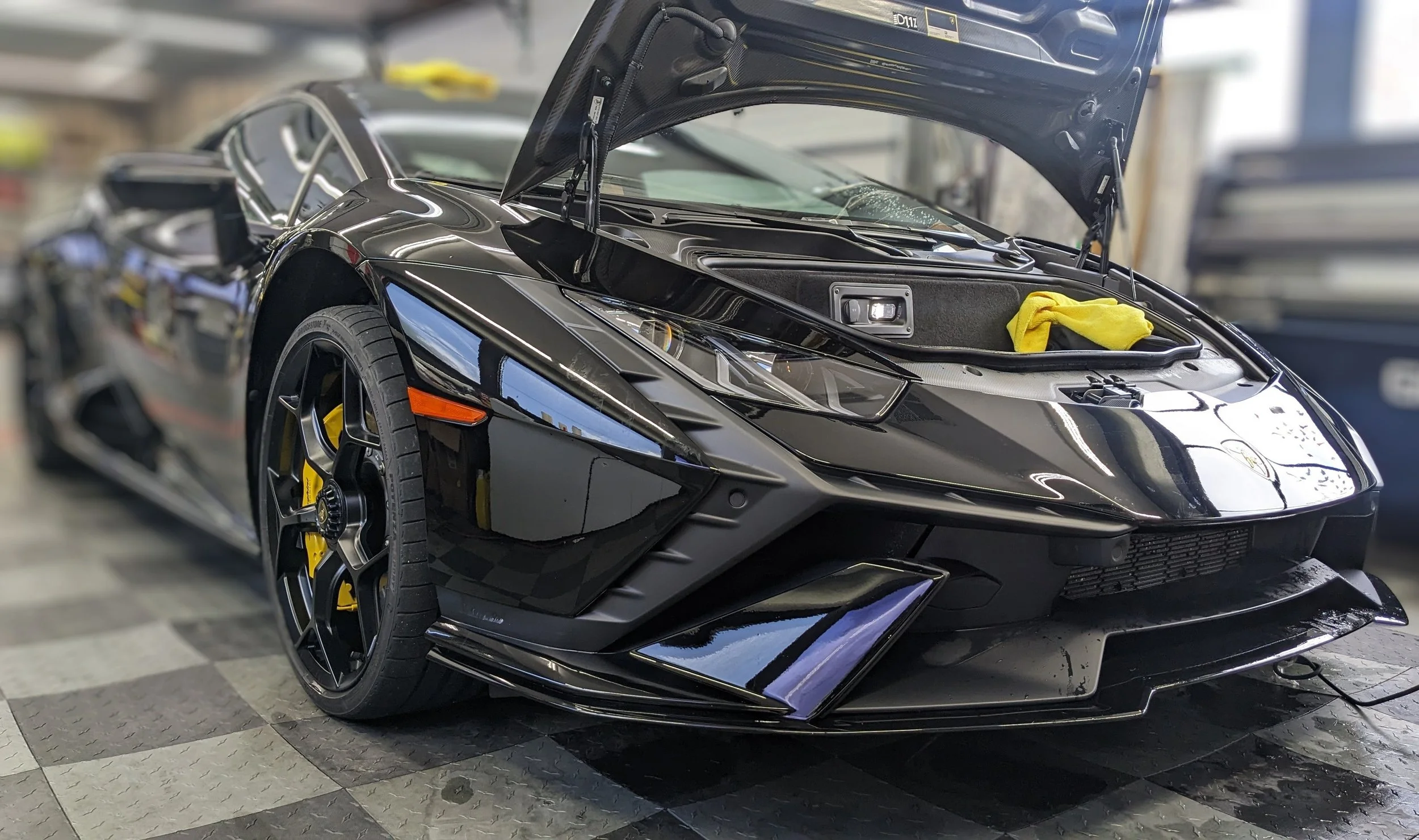 A black sports car with its hood open, showing the engine compartment, inside a garage or workshop with tools and equipment in the background.
