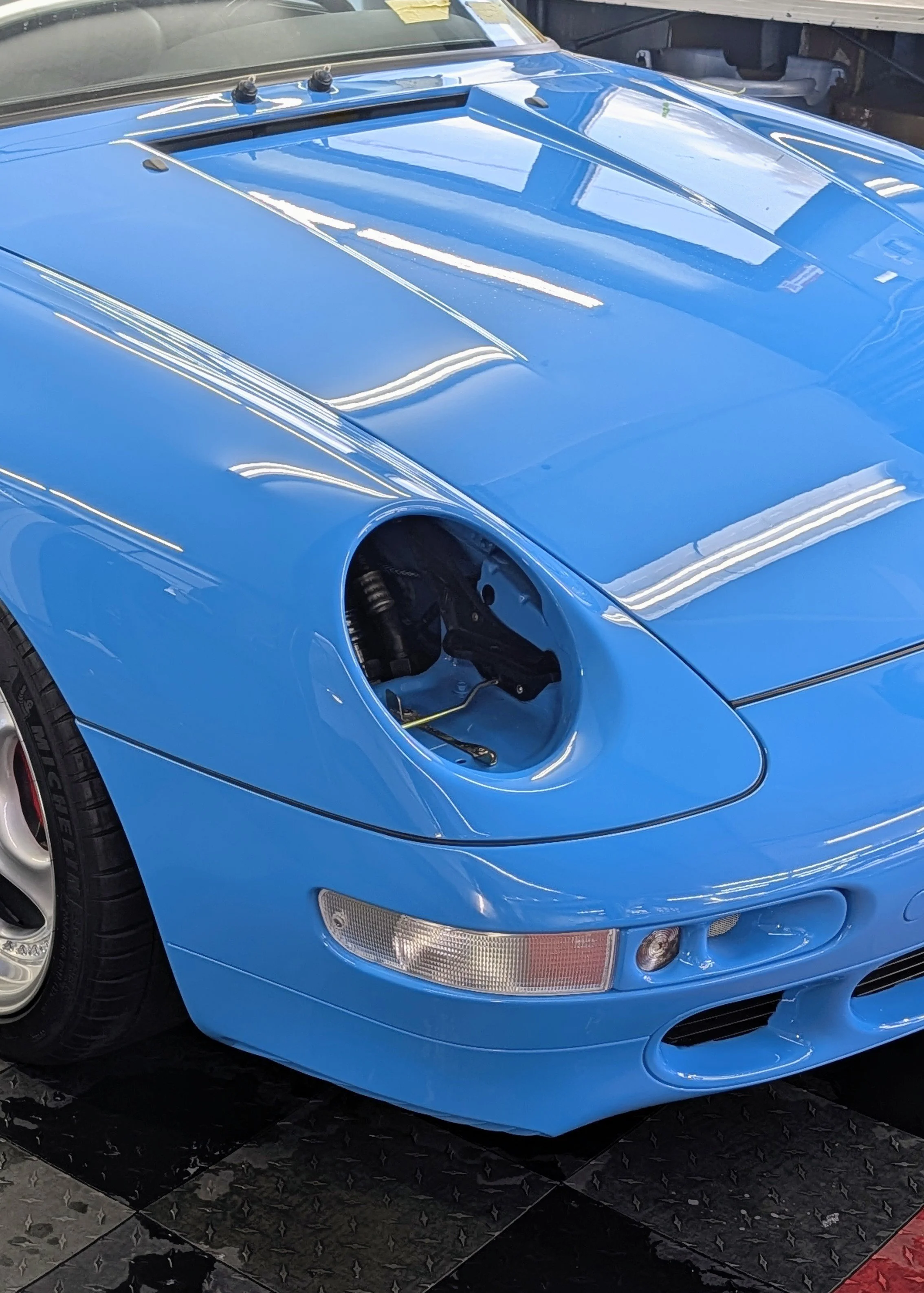 A blue sports car with an exposed headlight and no front bumper, showing the engine bay and wheel well.