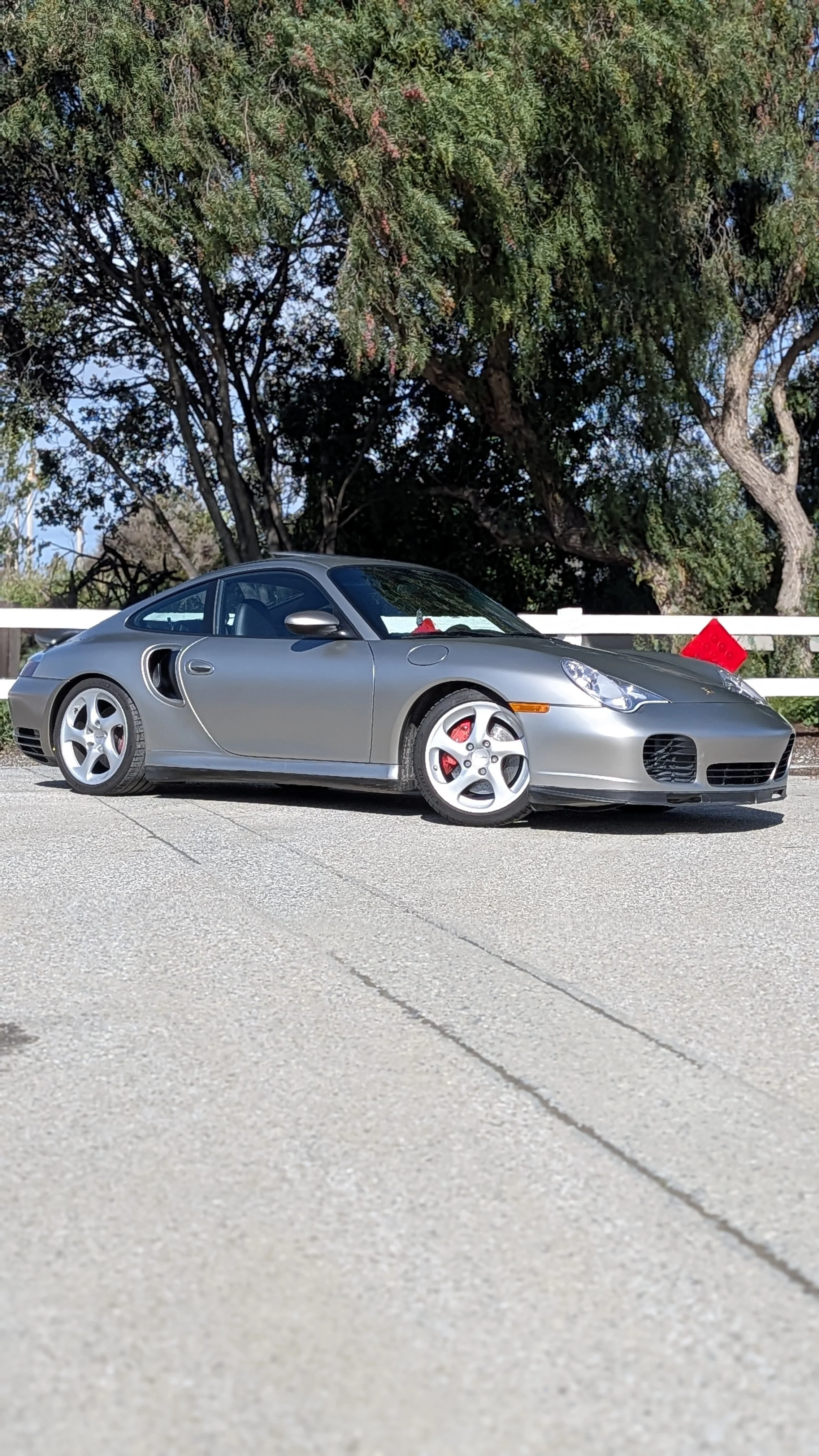 A silver Porsche 911 sports car parked on the side of the road, with a white fence and tall leafy trees in the background.