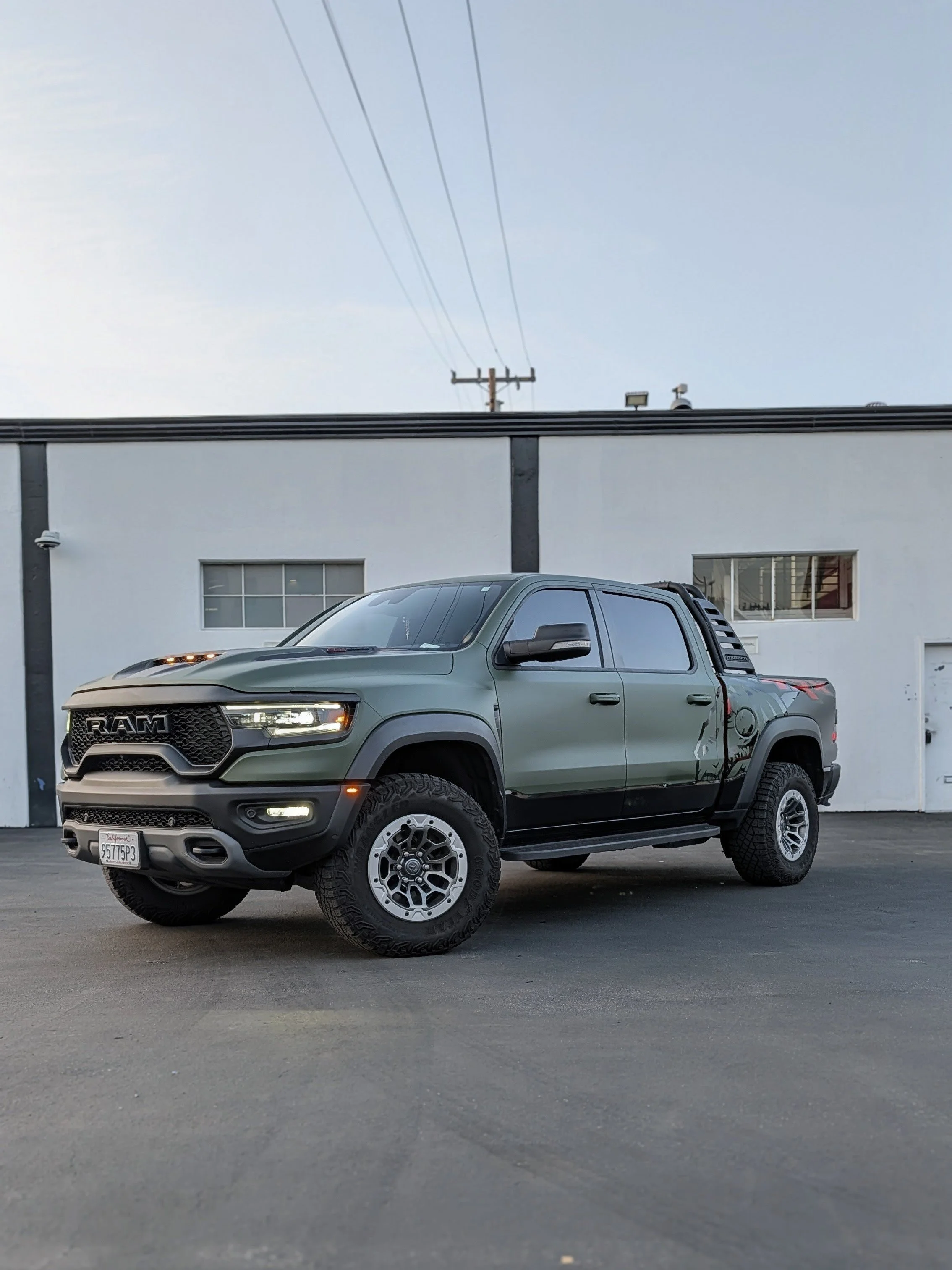 A green RAM pickup truck parked outdoors in front of a white industrial building with small windows and a closed garage door.