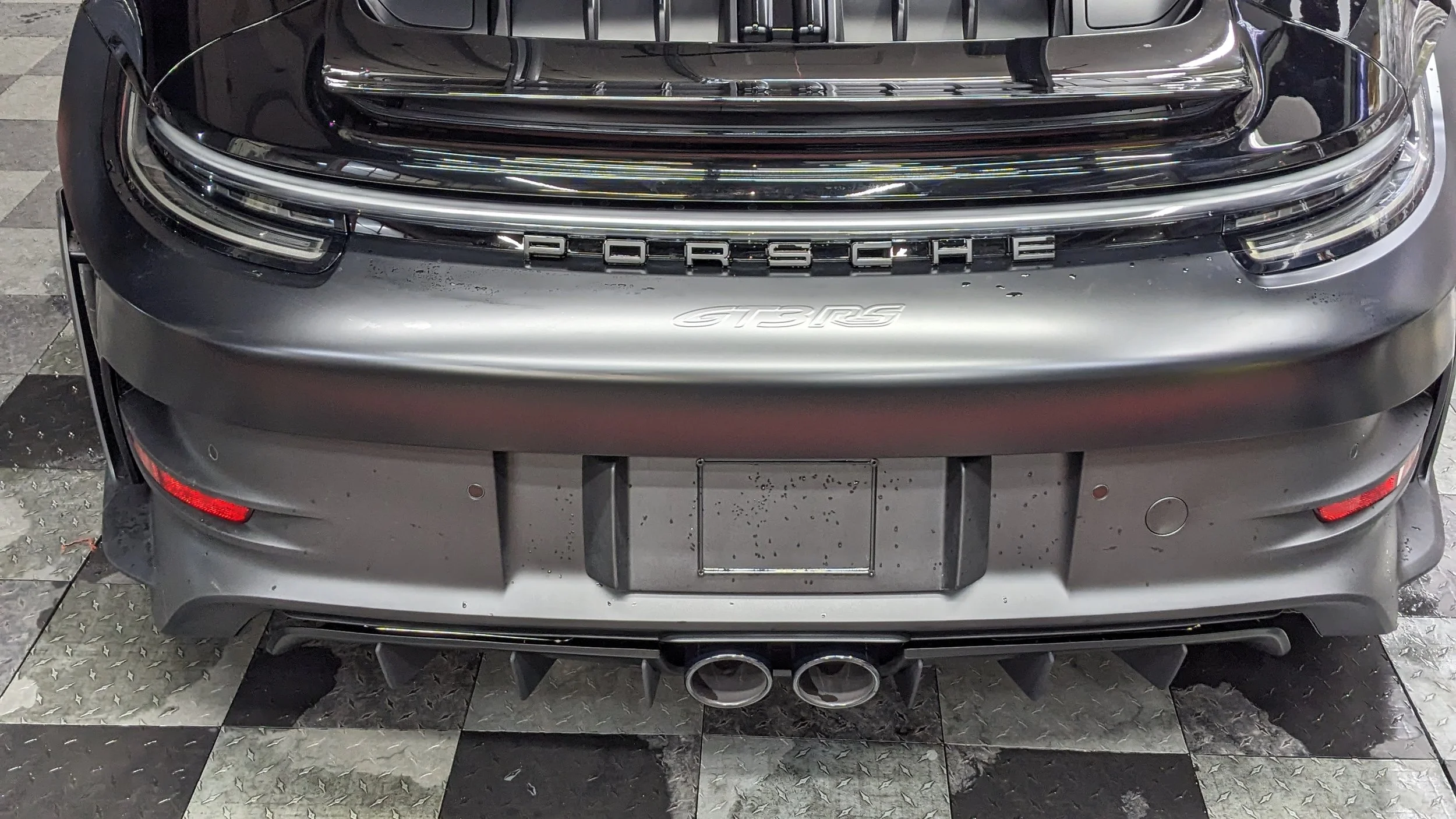 Rear view of a silver Porsche GT3 RS sports car parked on a checkered floor, showing dual exhaust pipes, rear bumper, taillights, and the Porsche emblem.
