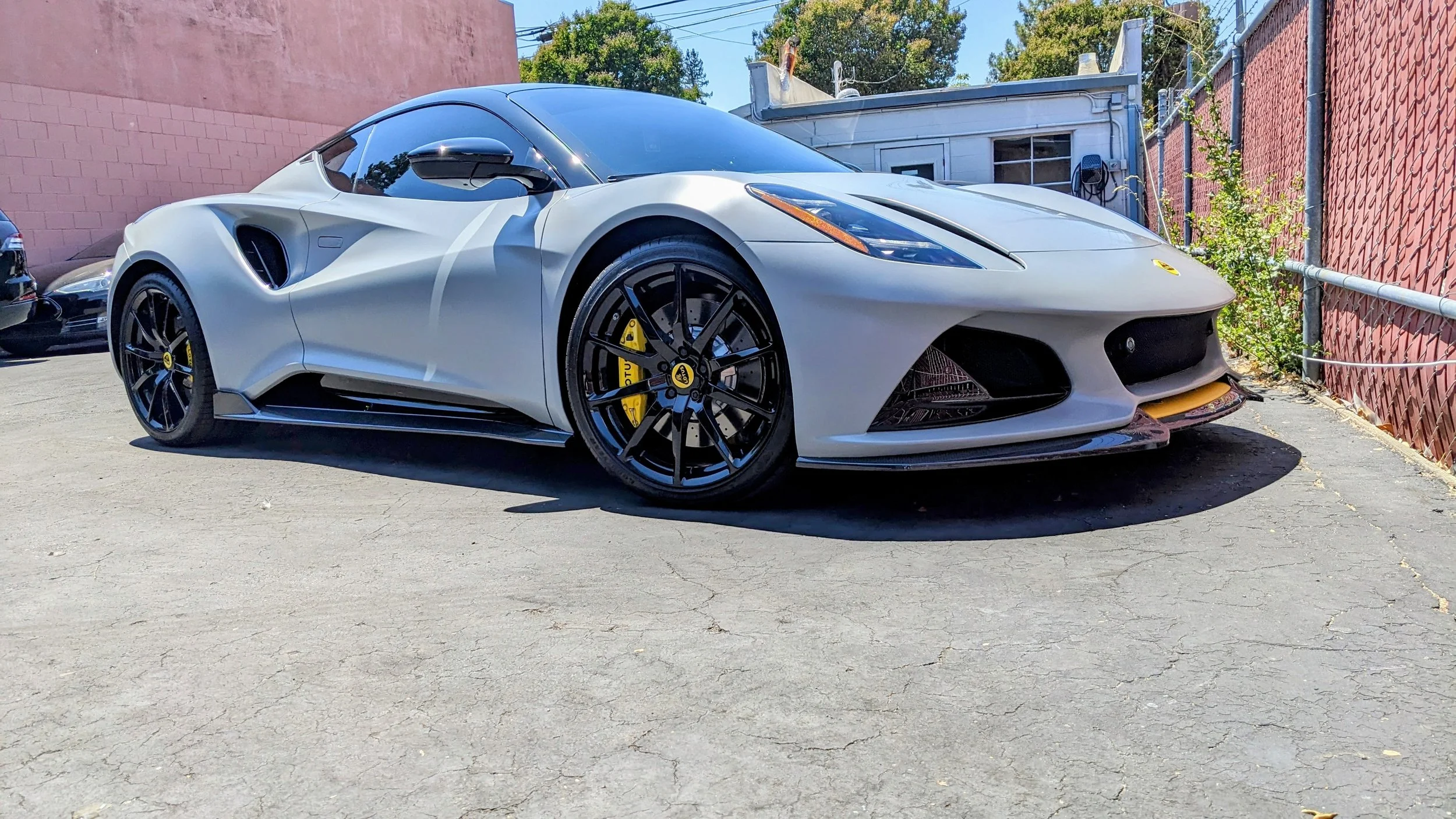 A silver sports car, a Lotus Evora, parked on a cracked asphalt surface next to a pink building and a red fence, with a white trailer in the background and some greenery.
