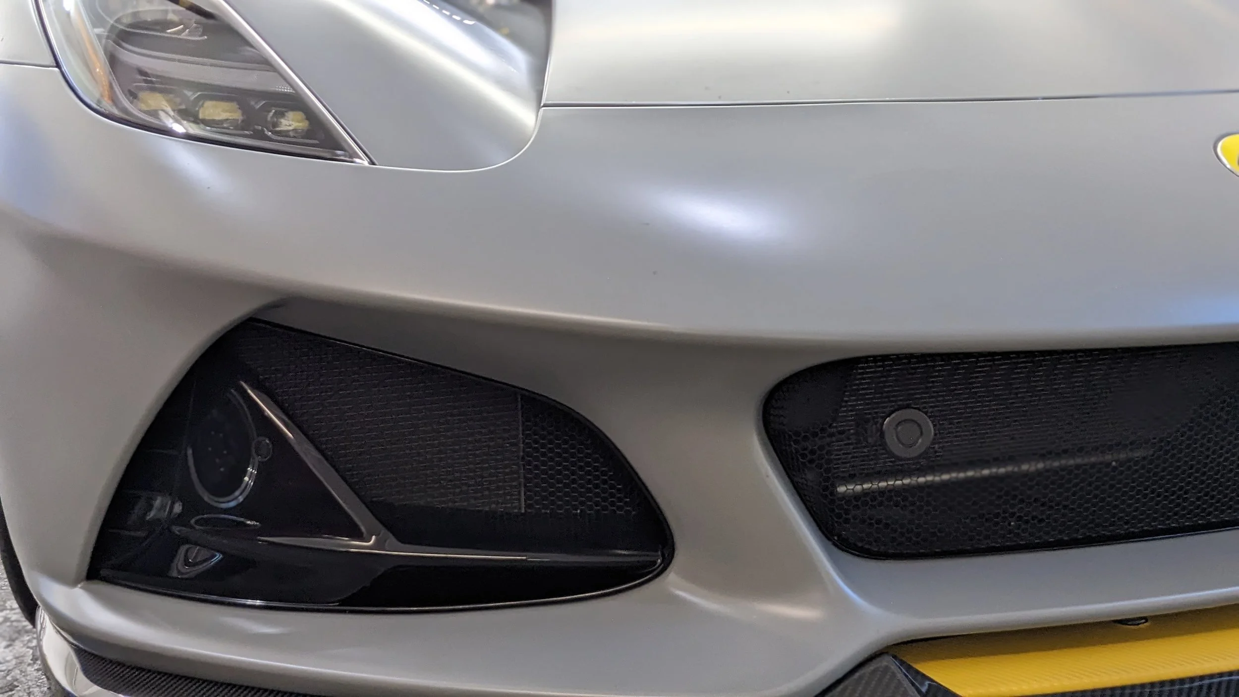 Close-up of the front end of a silver sports car, showing the headlight, front bumper, and air intakes.