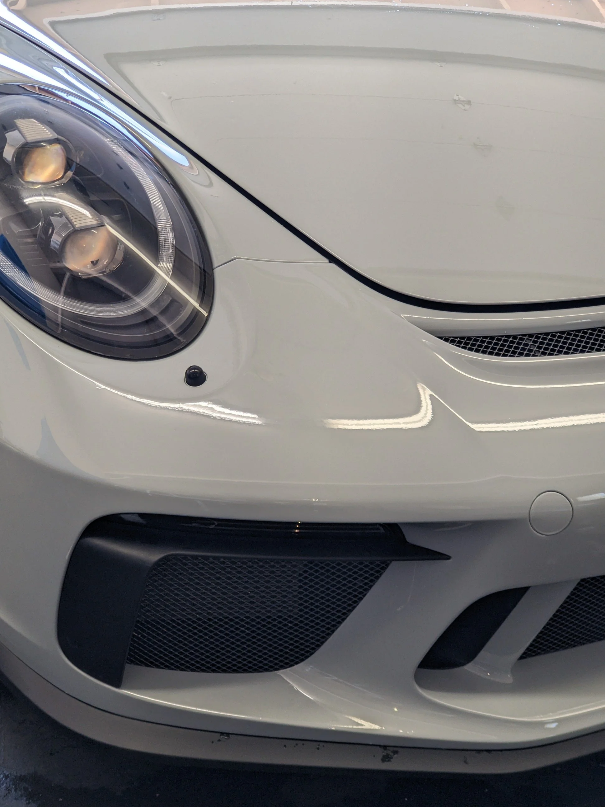 Close-up of the front of a white Porsche sports car, showing the headlight, front bumper, and grille.