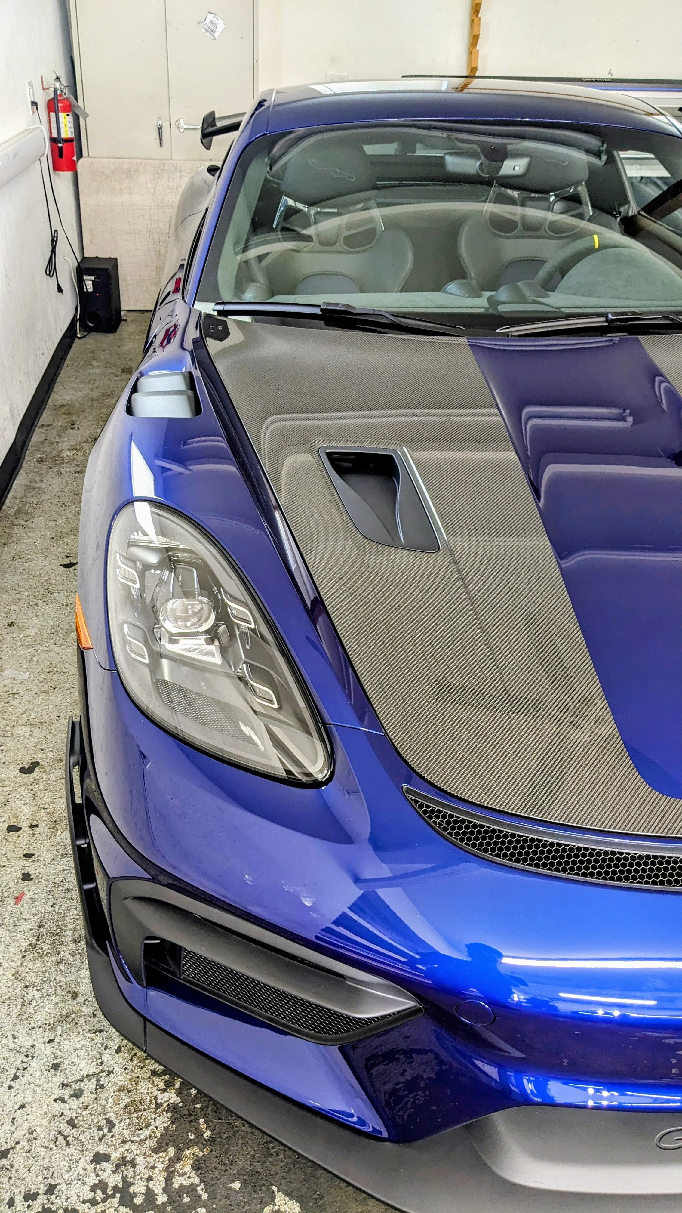 Blue sports car parked indoors, featuring a carbon fiber hood with a scoop and aerodynamic design.