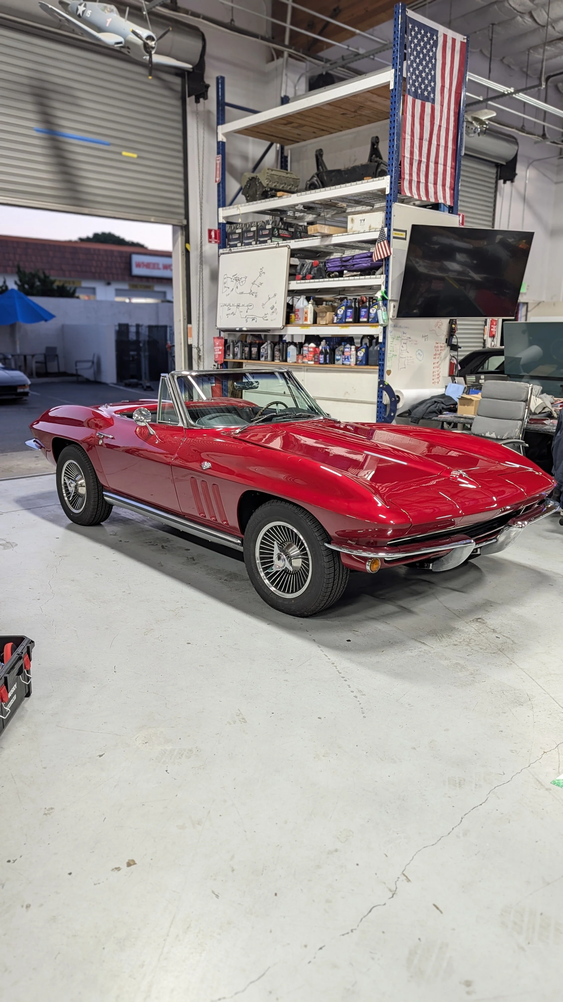 A red vintage convertible car inside a garage or workshop with American flags, a large TV, shelves of tools and parts, and a whiteboard with diagrams in the background.