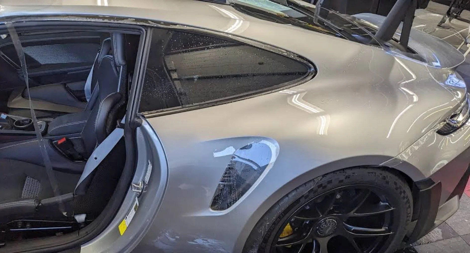 Silver sports car with black wheels and cockpit interior, raindrops on windows, and a partial view of the front and side.
