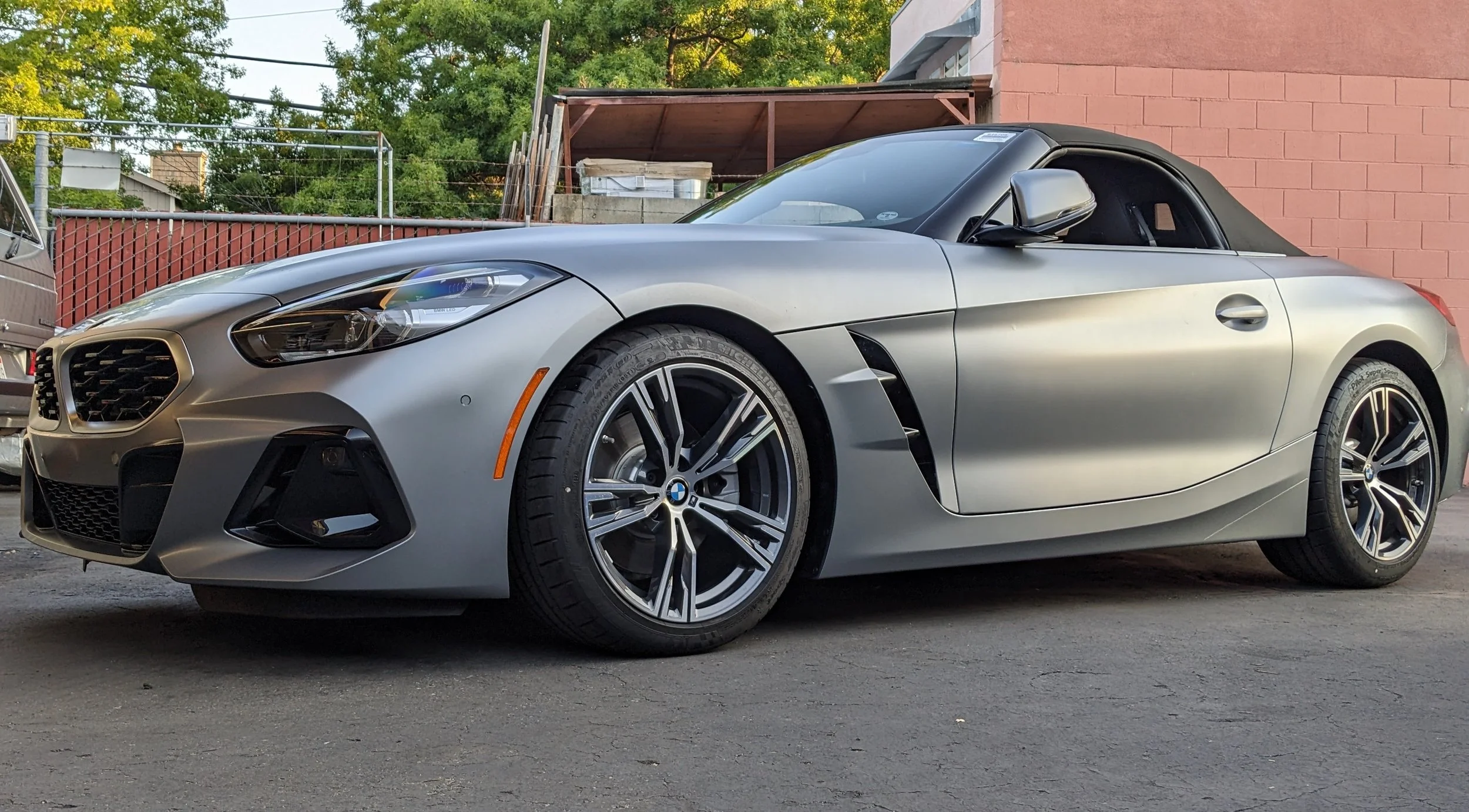 A silver BMW convertible sports car parked on an asphalt surface in front of a red brick building and a wooden fence with green trees in the background.