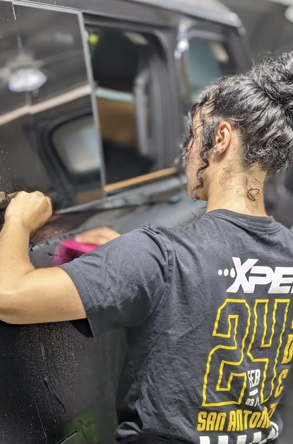 A woman cleaning a black vehicle's window with a pink cloth in a garage or workshop.