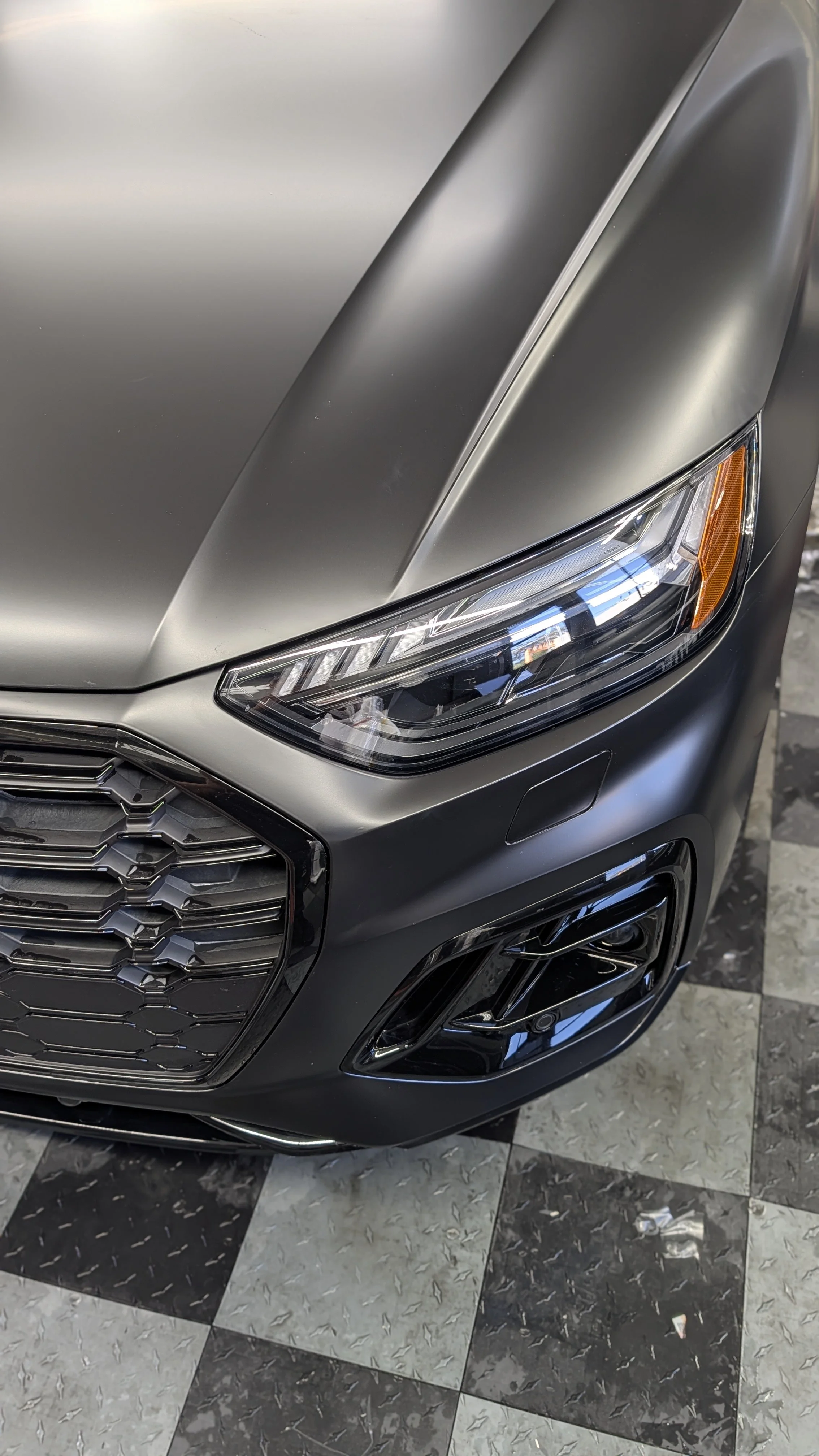 Close-up of the front part of a gray sedan car from a high angle, showing the headlights, grille, and part of the hood, parked on a checkered floor.