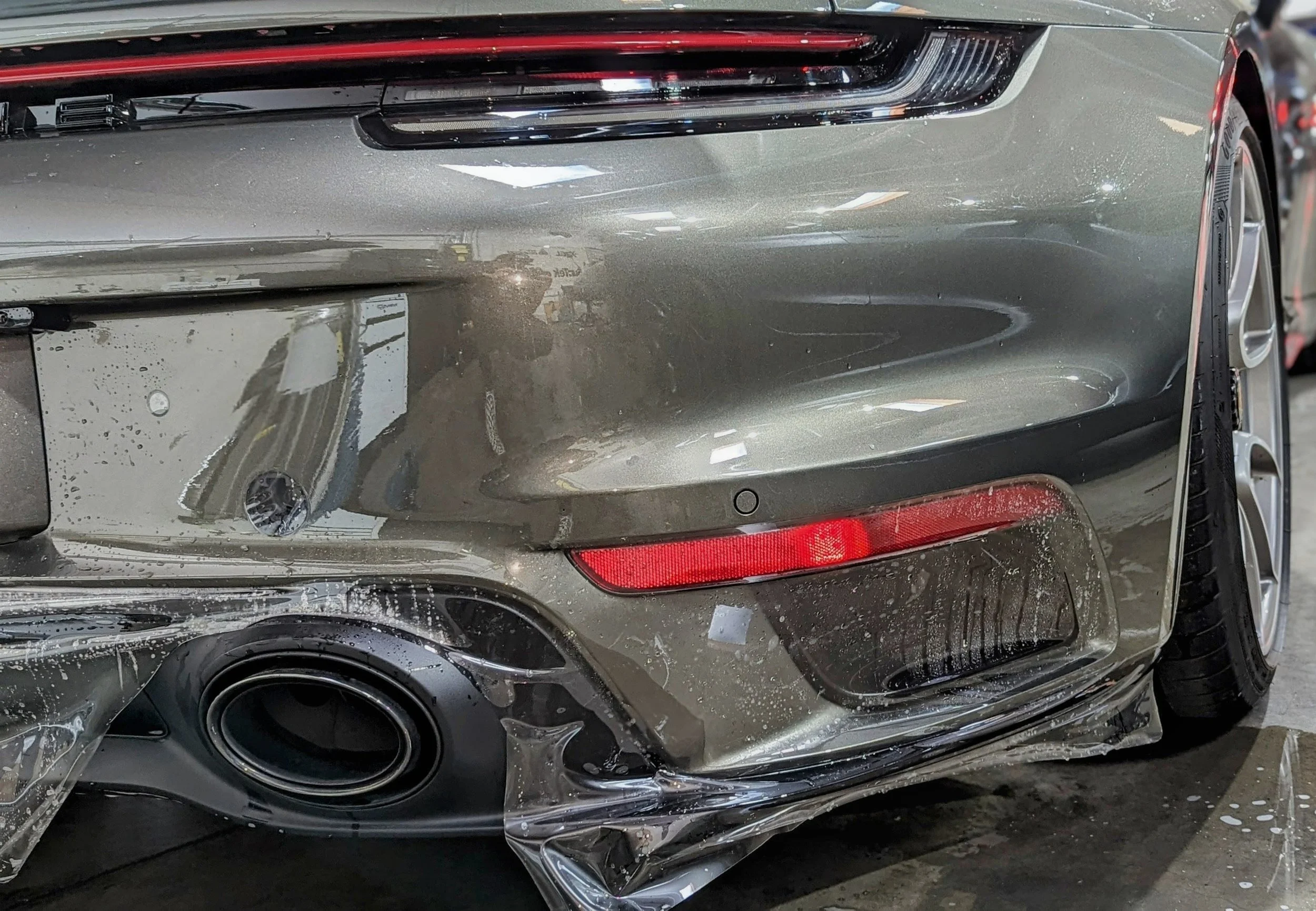 Close-up of a silver luxury sports car with front bumper damage, a red reflector, and visible scratches and dents.