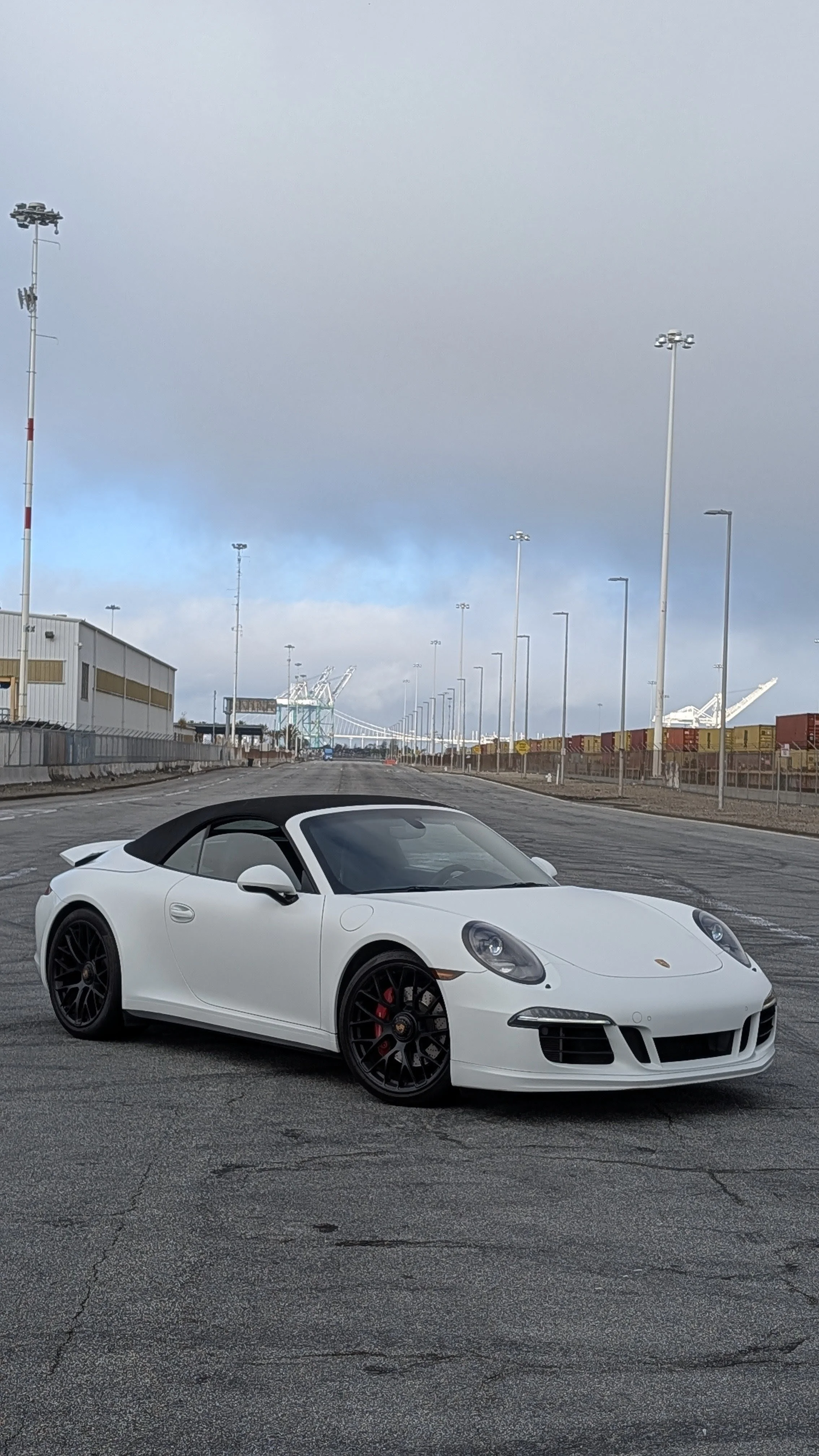 A white Porsche convertible sports car with a black soft top parked on an empty industrial lot, with shipping cranes and containers visible in the background under cloudy skies.