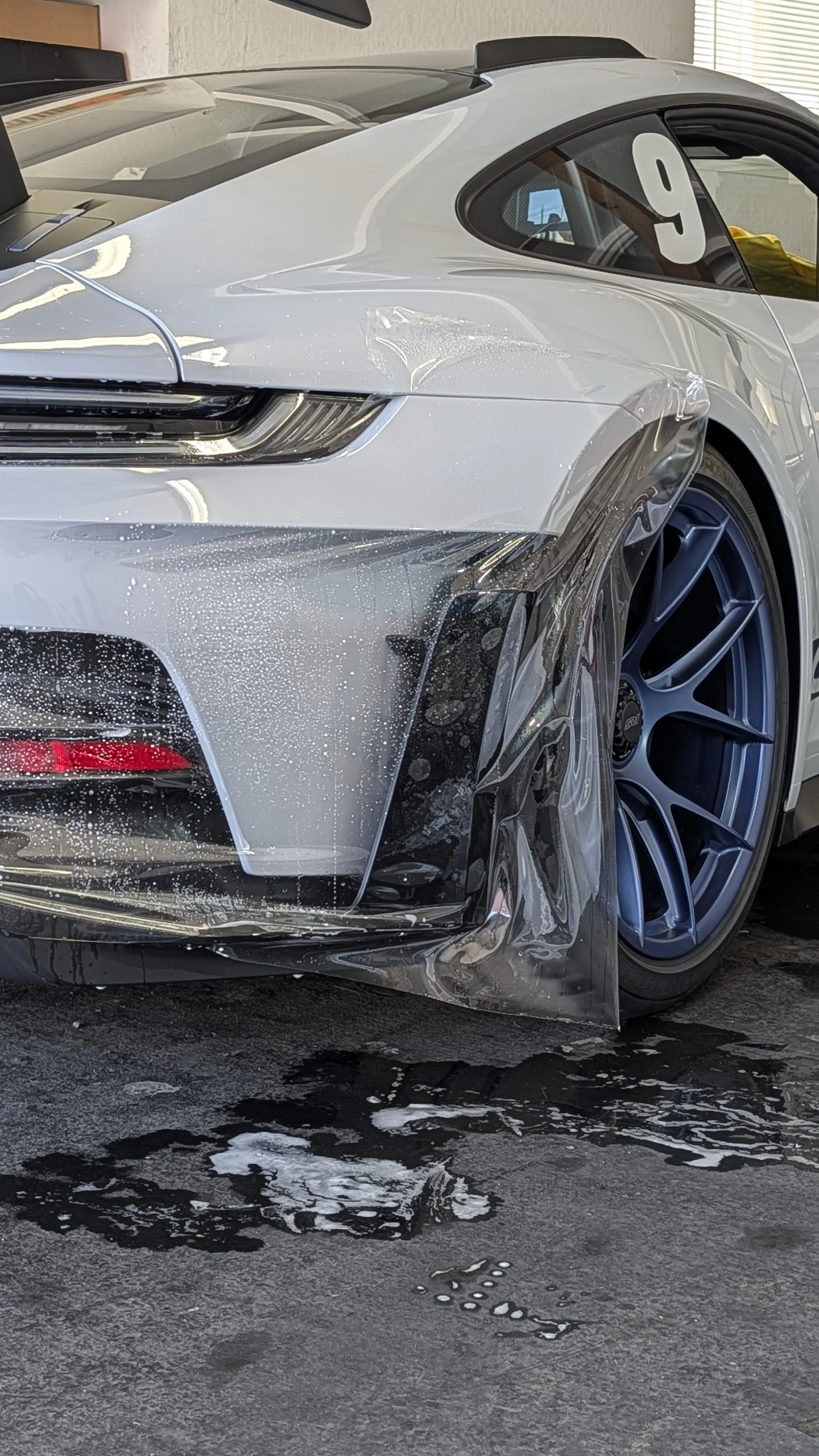Close-up of a silver sports car with damage to the front left side, covered in soap suds, in a cleaning or detailing process.