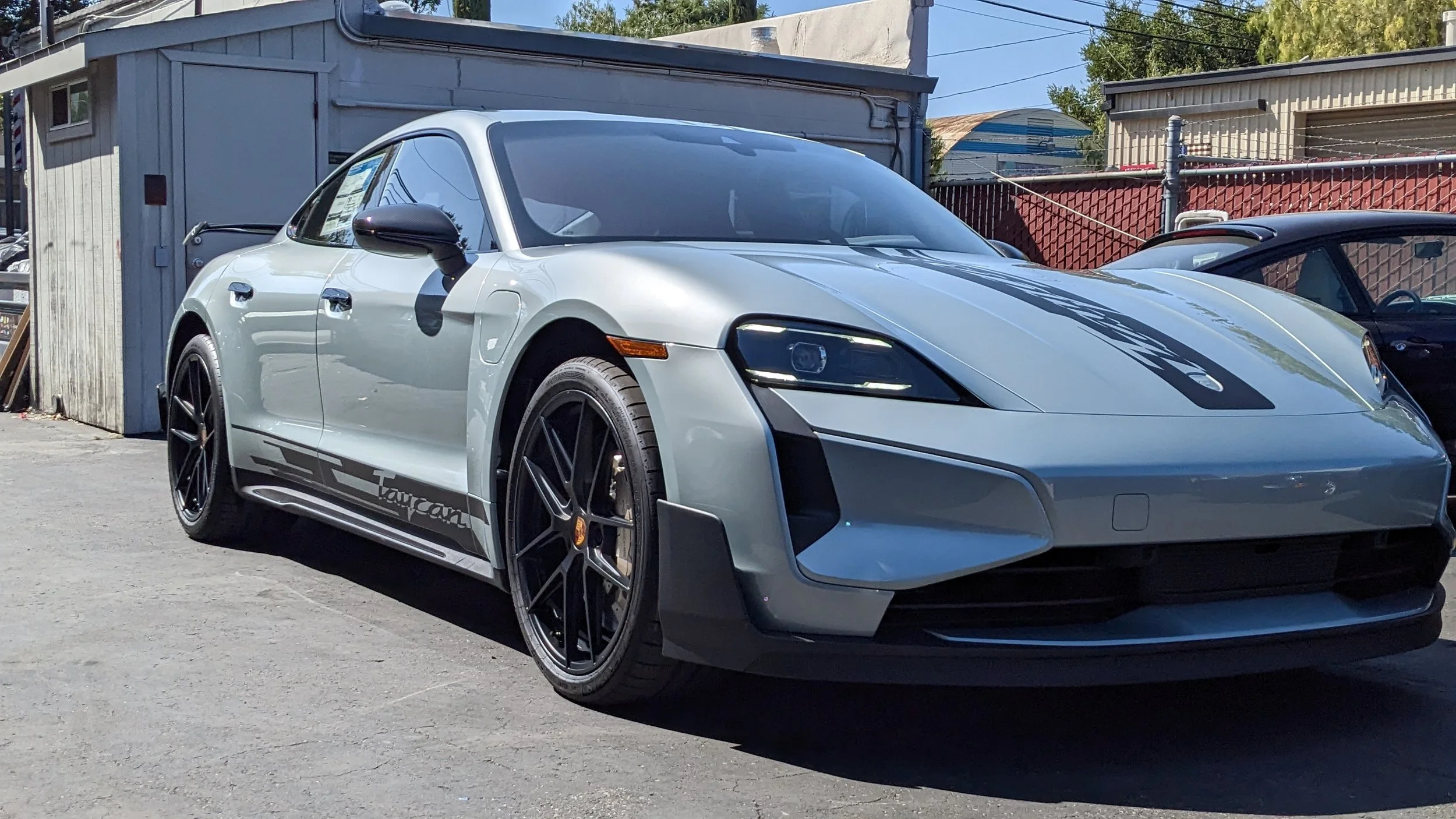 A silver Porsche Taycan sports car parked outdoors on a paved surface, with black wheels, black racing stripes on the hood, and black side mirrors, next to a small building.