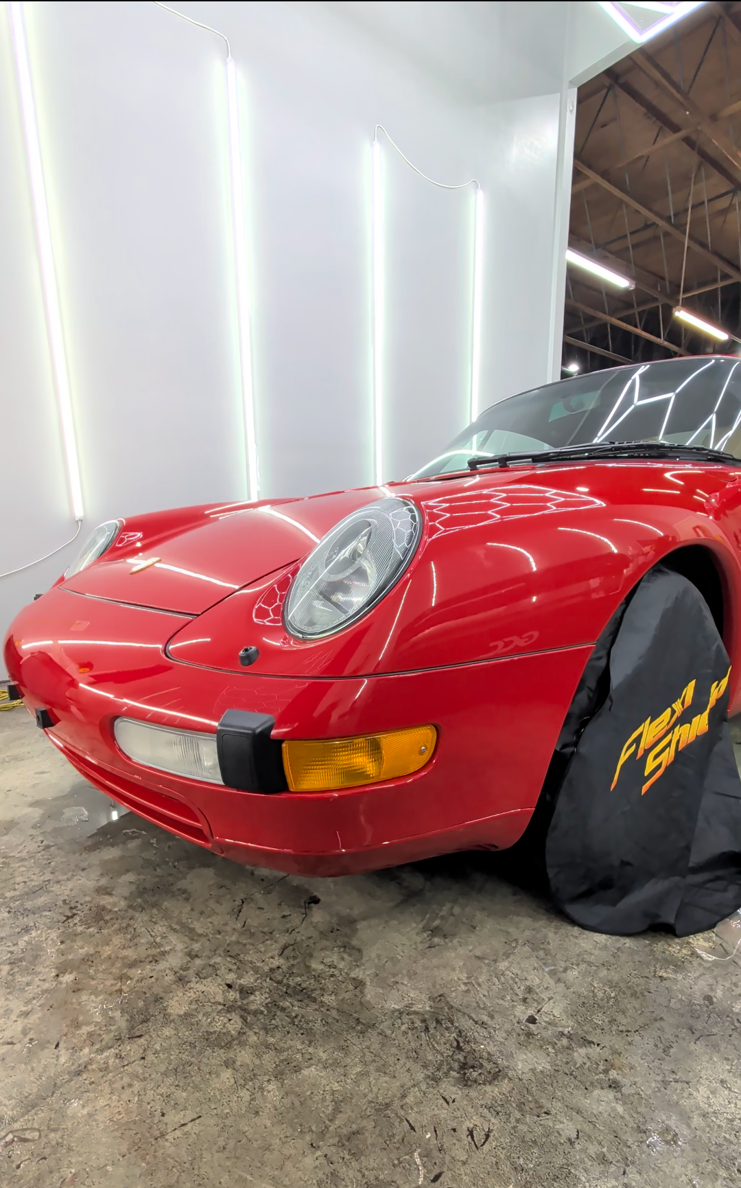 Red classic sports car parked in a garage with LED strip lights on the wall, one wheel covered with a black tire cover labeled 'Next Gen'.