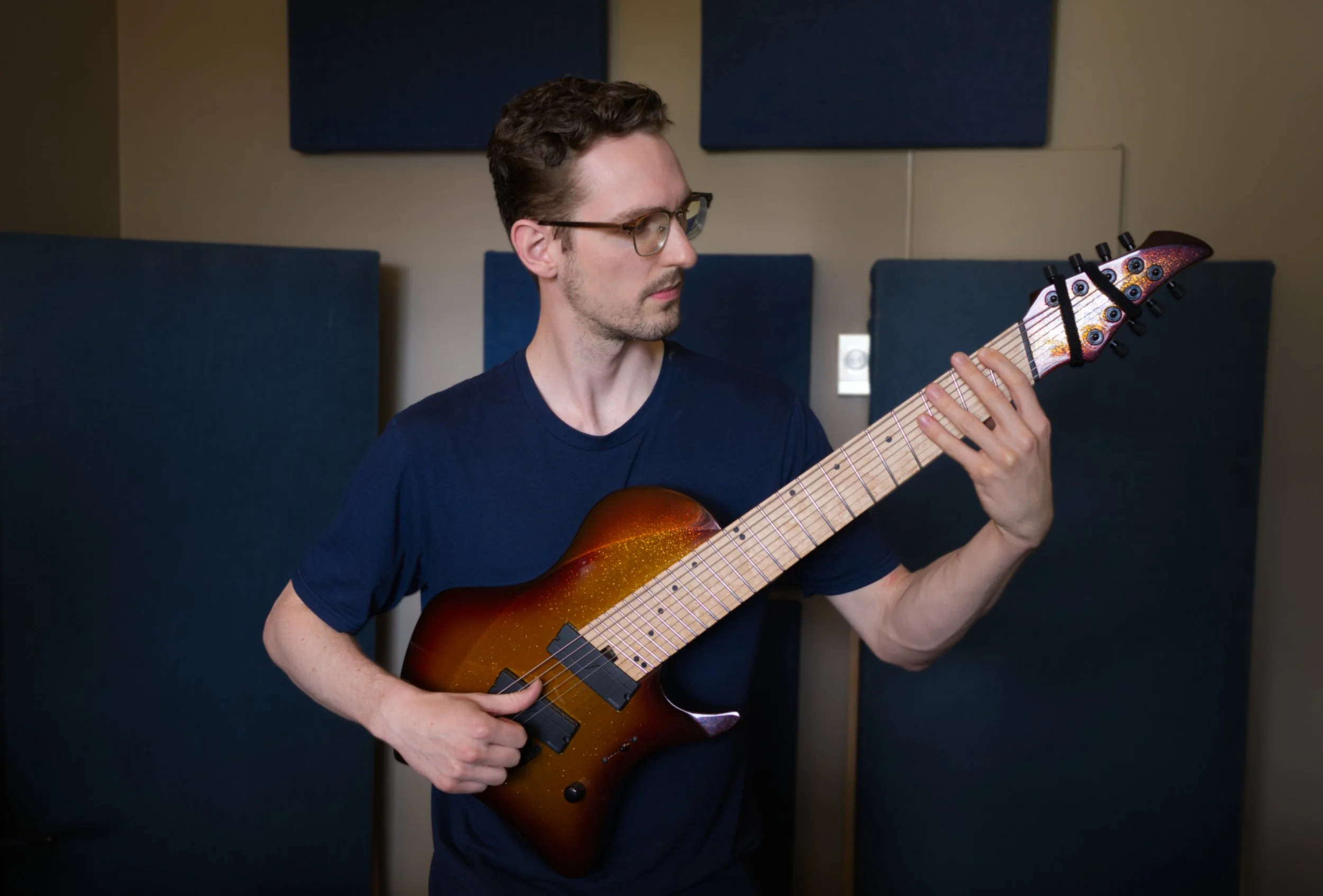 A young man with glasses and dark hair, playing an 8-string electric guitar in a home music studio.