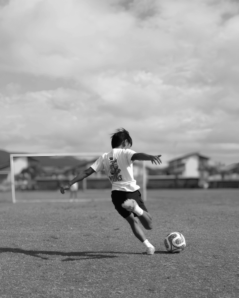 A boy playing soccer on a grassy field during the daytime, with goalposts and houses in the background, under a partly cloudy sky.