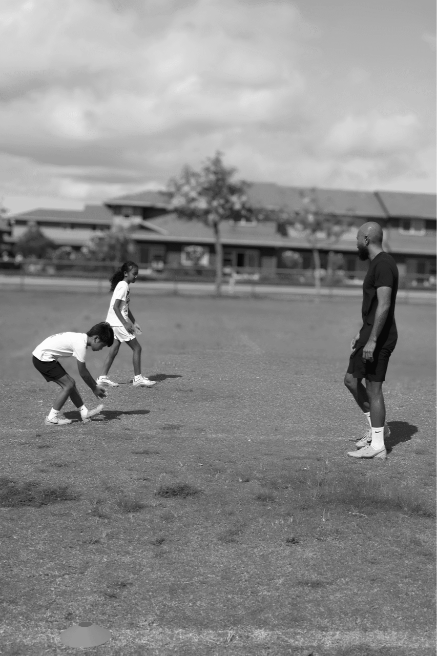 A man and two children playing a game on a grassy field outdoors, with houses and trees in the background and cloudy sky overhead.