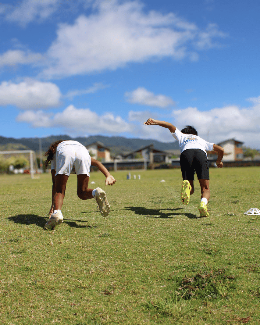 Two children running on a grassy field during a sunny day, with houses and mountains in the background.