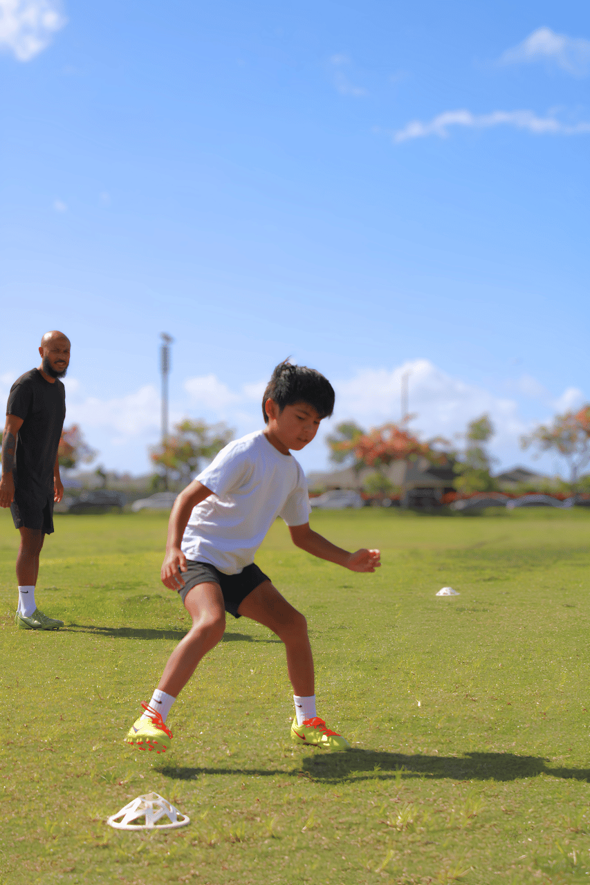 A boy practicing soccer drills on a grassy field, with a coach watching in the background during daytime.