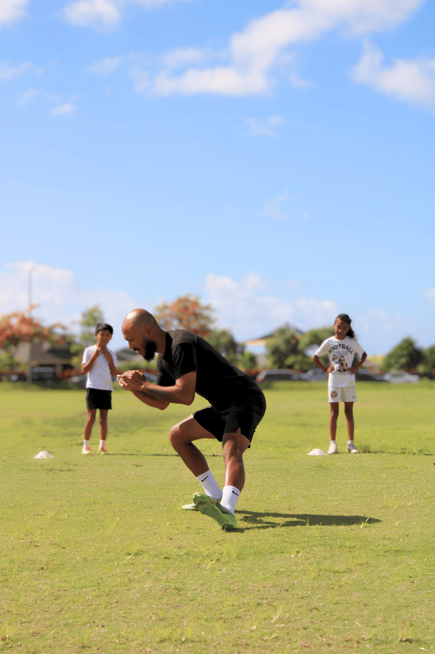 A man in athletic wear crouching on a grassy field, with two children watching in the background on a sunny day.