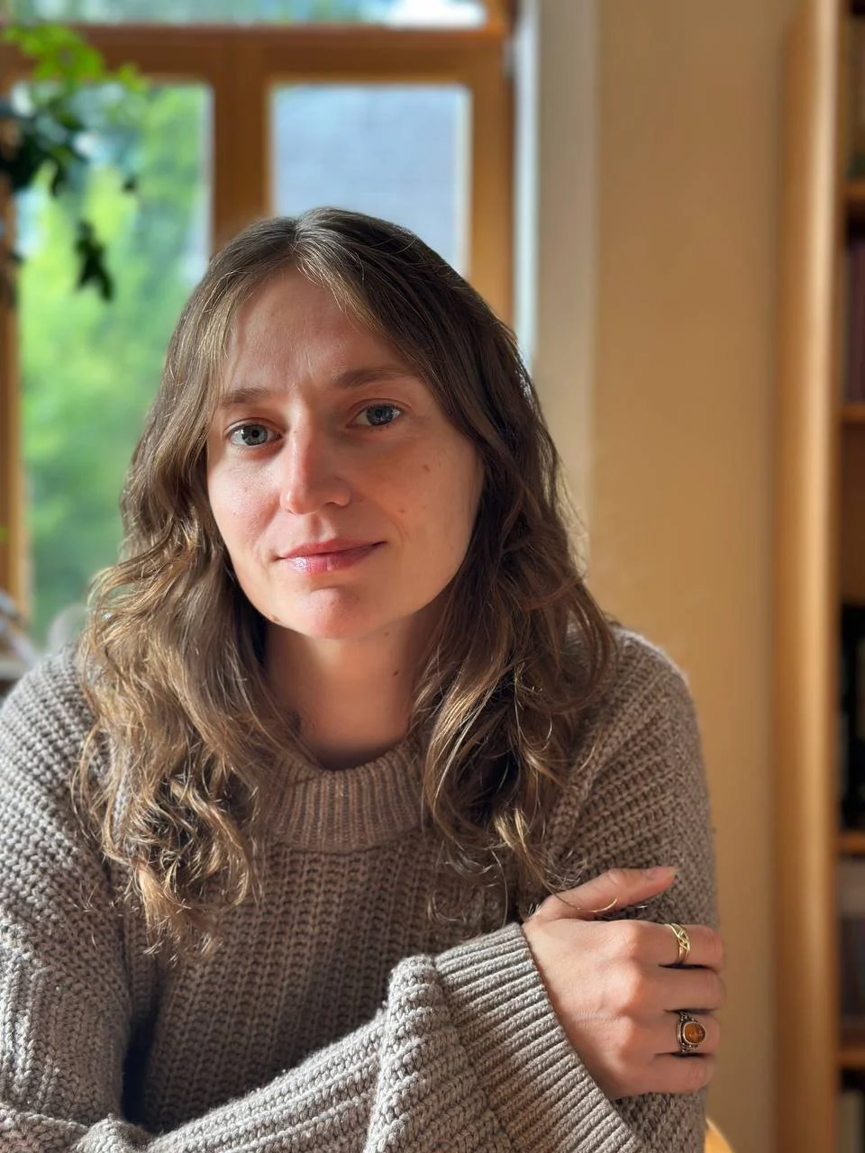 Junge Frau mit braunen, lockigen Haaren und blauen Augen, trägt einen beige Strickpullover, sitzt in einem Zimmer mit Schaukelstuhl, Fenster im Hintergrund, blickt in die Kamera.