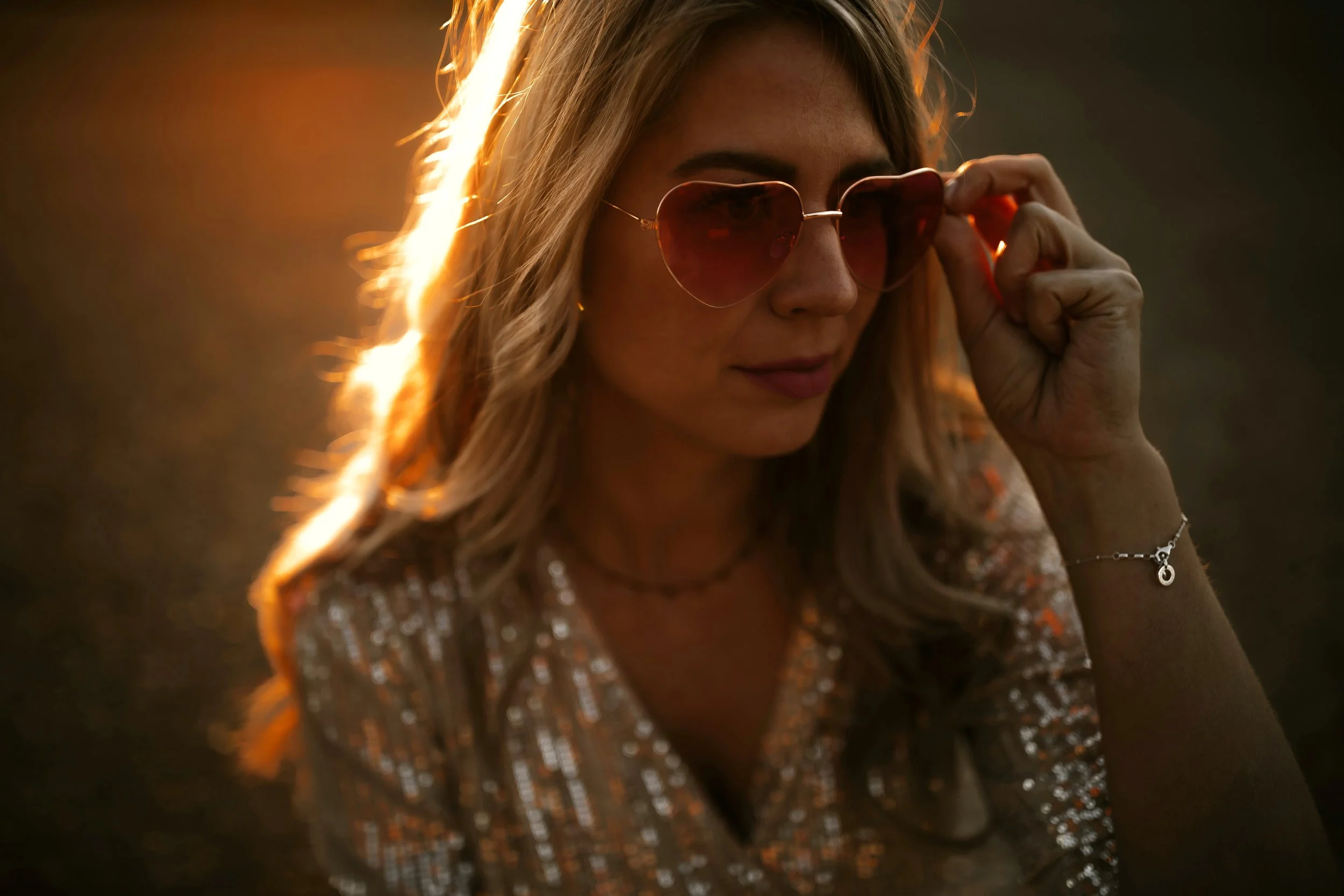 Junge Frau mit Sonnenbrille vor einem Feuer, trägt ein glitzerndes Top und hat blonde, lockige Haare. Lustige Romcom Autorin Roosa Root