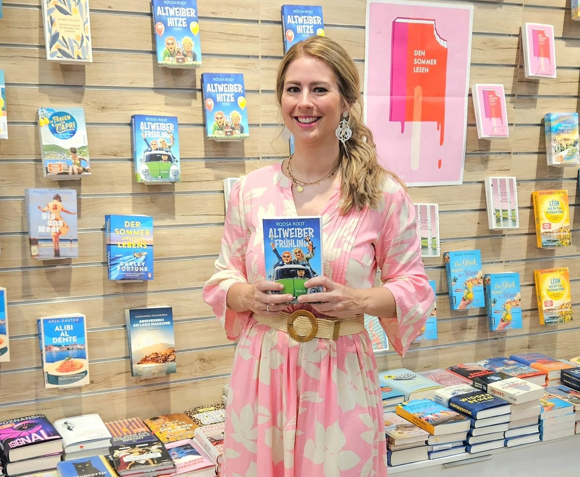 Frau Roosa Root, Autorin mit Buch in der Hand Altweiber-Hitze. Vor Bücherwand Rosa Kleid