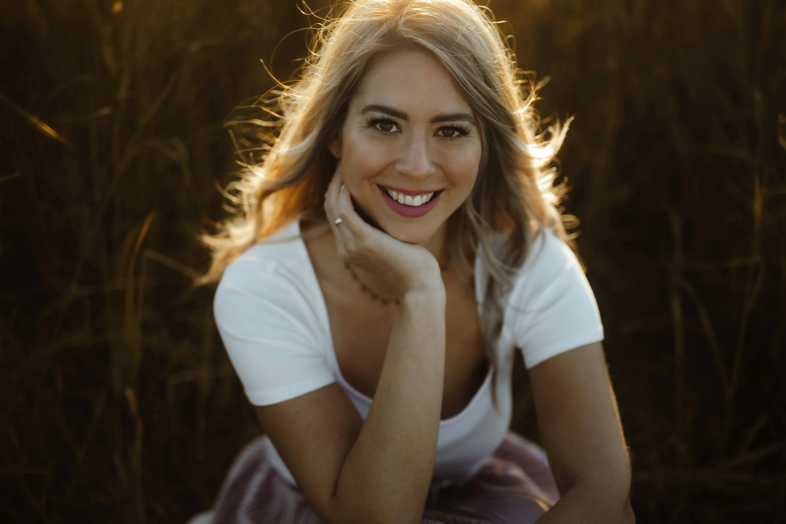Eine junge Frau mit langem, hellblondem Haar lächelnd, sitzt im Freien bei Sonnenuntergang in einem Feld. Sie trägt ein weißes T-Shirt und ist in einer entspannten Pose. Der Hintergrund ist leicht verschwommen, mit warmen, goldenen Tönen vom Sonnenli