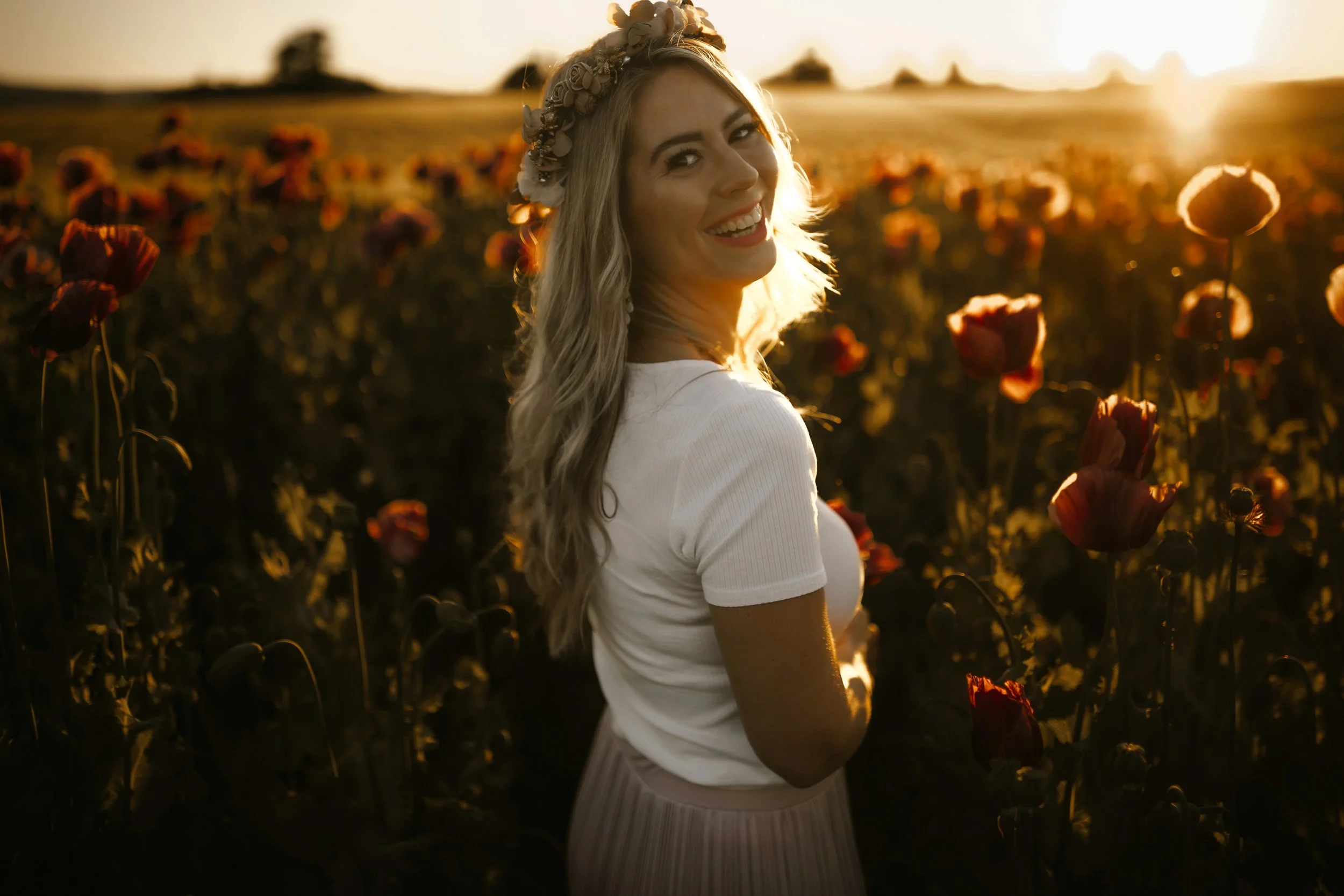 Eine lachende Frau mit langem, lockigem Haar trägt einen Blumenkranz und ein weißes T-Shirt, steht in einem Feld mit roten Mohnblumen bei Sonnenuntergang.