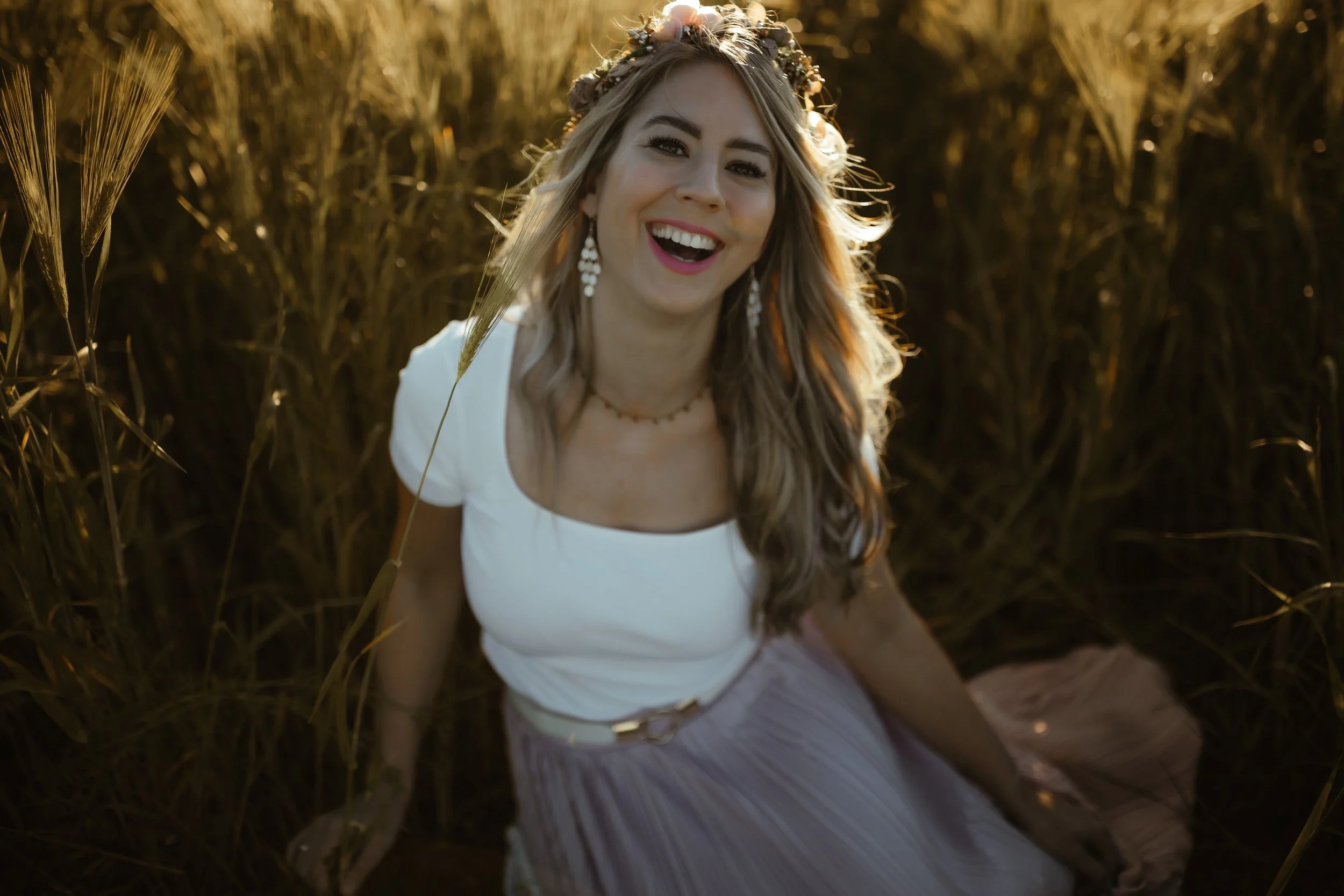 Eine lachende junge Frau mit langen, gewellten Haaren sitzt im Feld bei Sonnenuntergang, trägt ein weißes Oberteil, einen Blumenkranz im Haar und große, hängende Ohrringe.