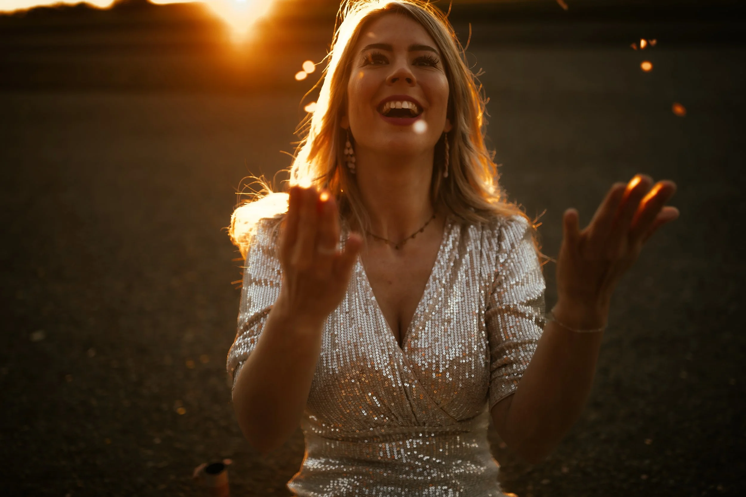 Junge Frau im glitzernden Kleid im Sonnenuntergang, die nach oben greift und auf den Boden schaut, während Lichtreflexe um sie herum leuchten.