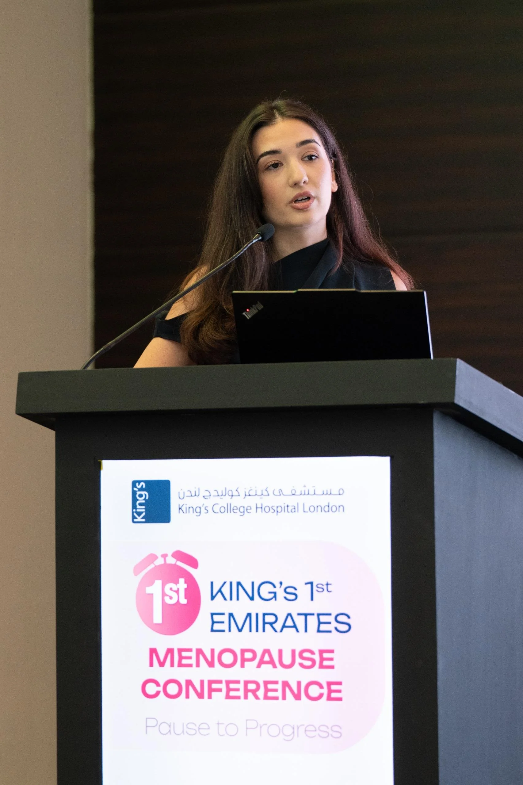 A woman standing at a podium giving a presentation at the King’s 1st Emirates Menopause Conference, with a laptop open in front of her and a sign on the podium display attend a healthcare conference.