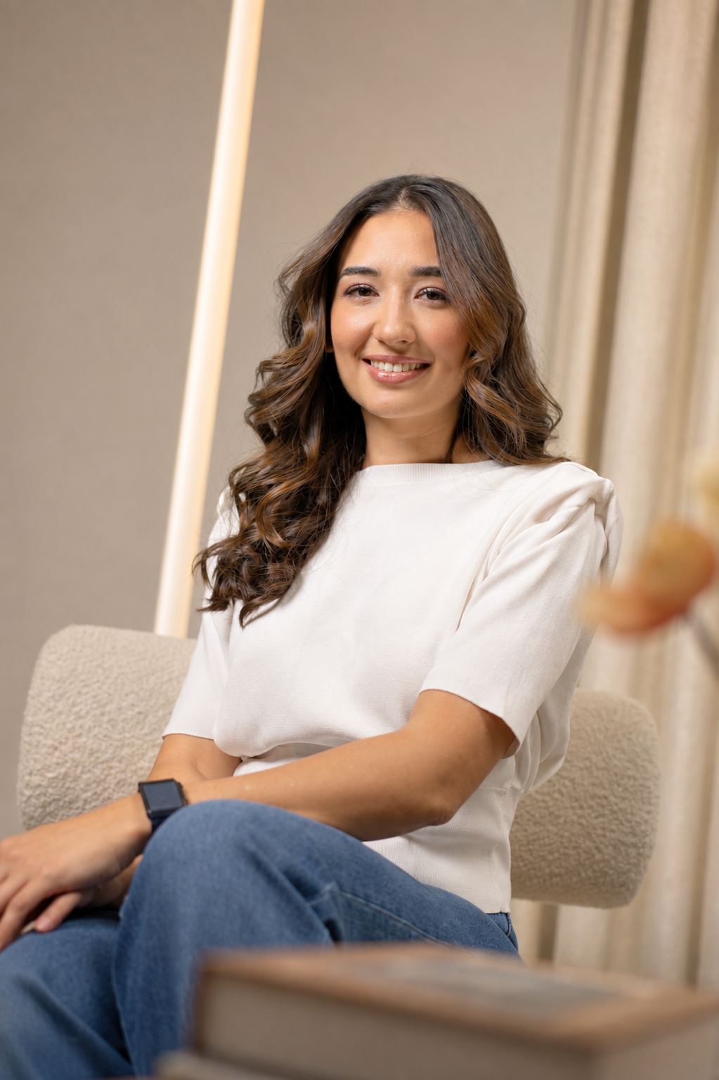 A young woman with long, wavy brown hair smiling and sitting on a beige chair indoors. She is wearing a white t-shirt and blue jeans, with a black watch on her left wrist. There are beige curtains and a gray wall in the background.