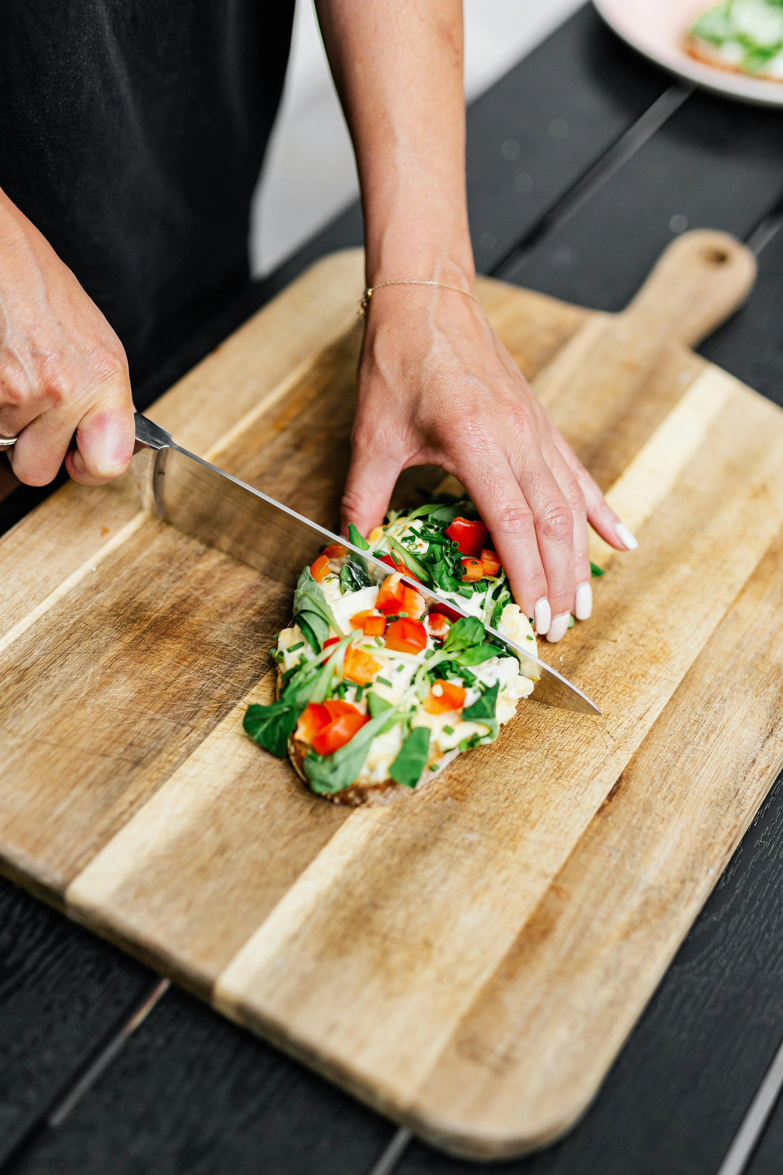 A person slicing a piece of bread topped with vegetables on a wooden cutting board.