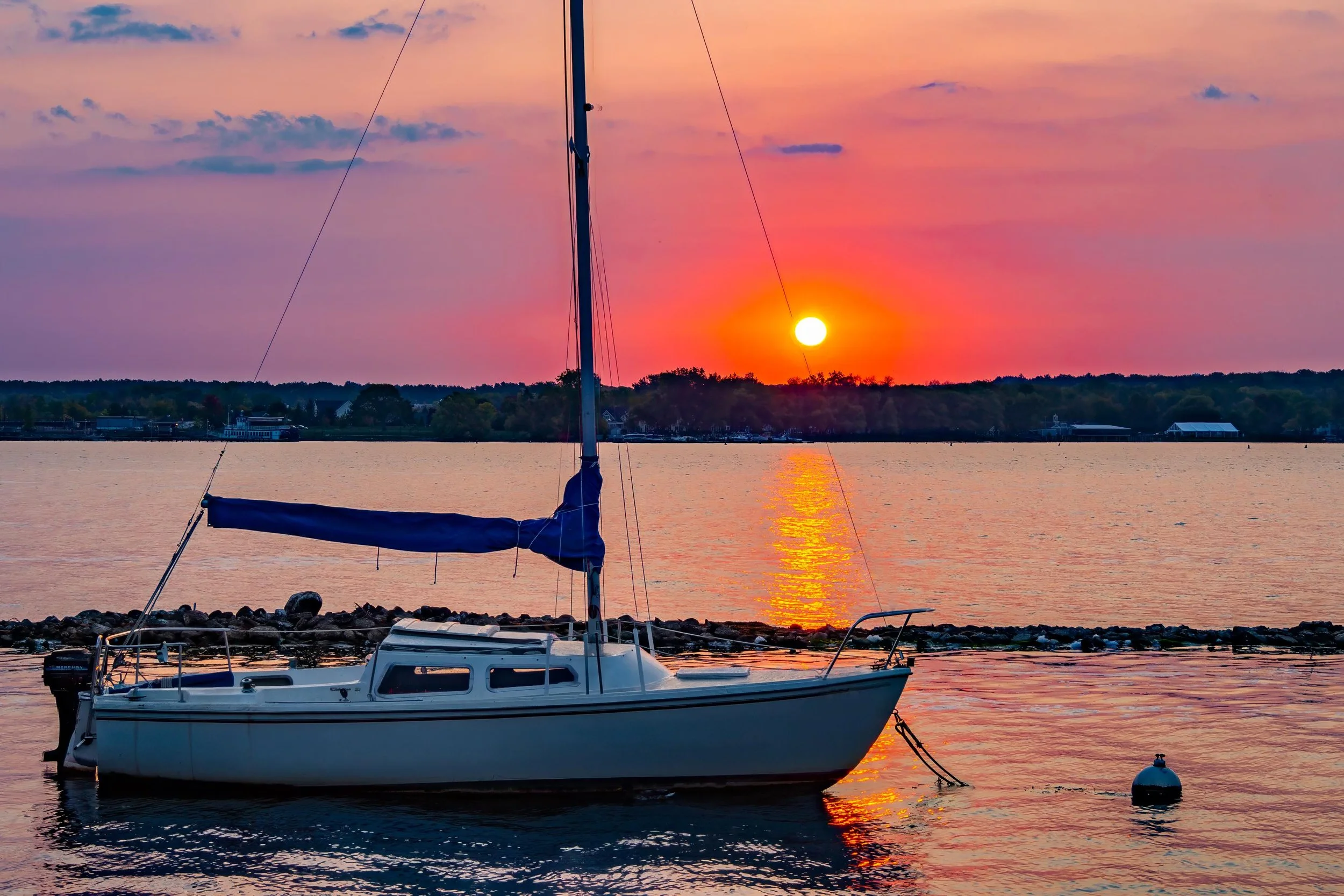A sailboat anchored in a body of water during sunset, with the sun near the horizon and colorful sky reflected on the water.